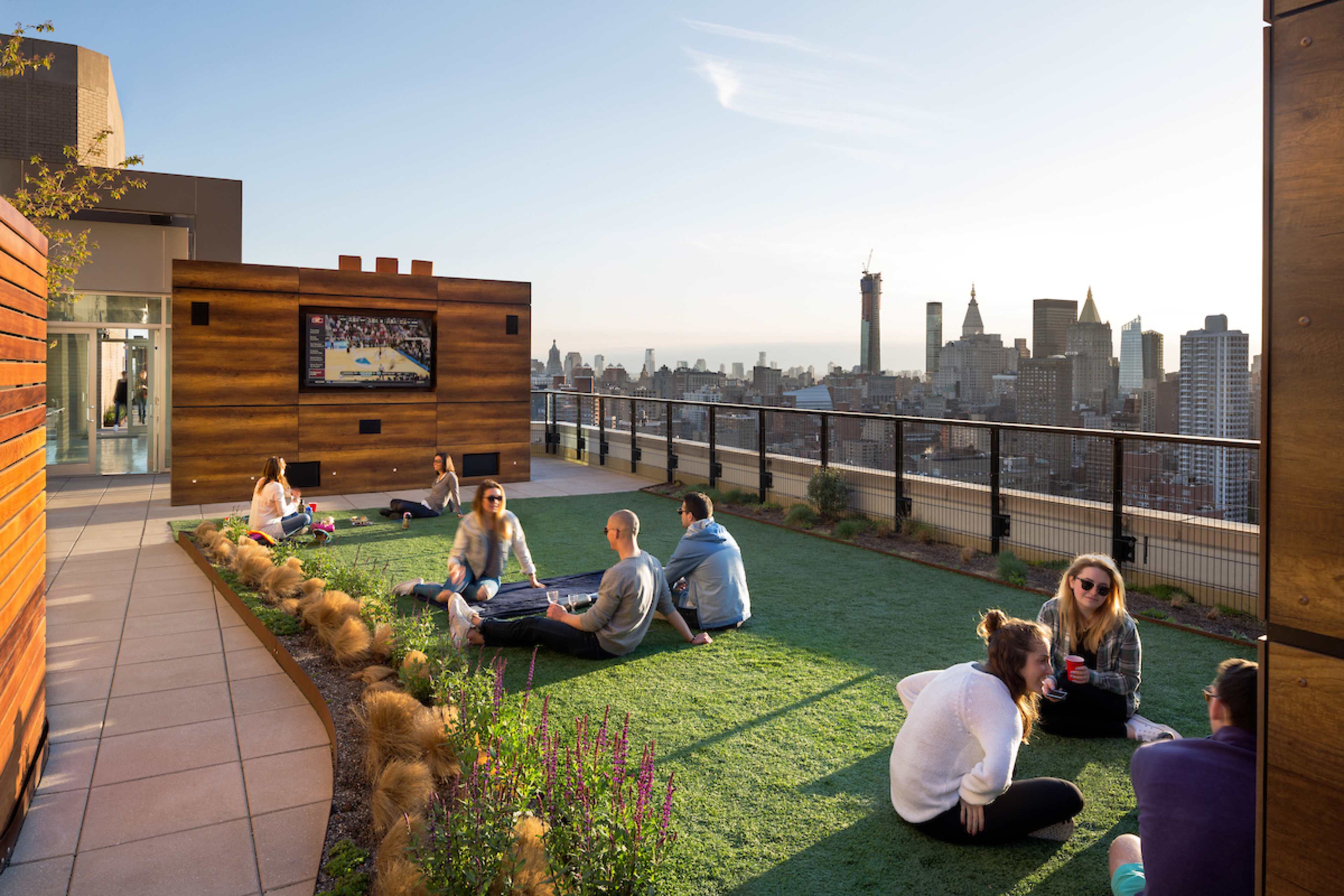 People relax on a green rooftop space with a view of a city skyline and a television display.