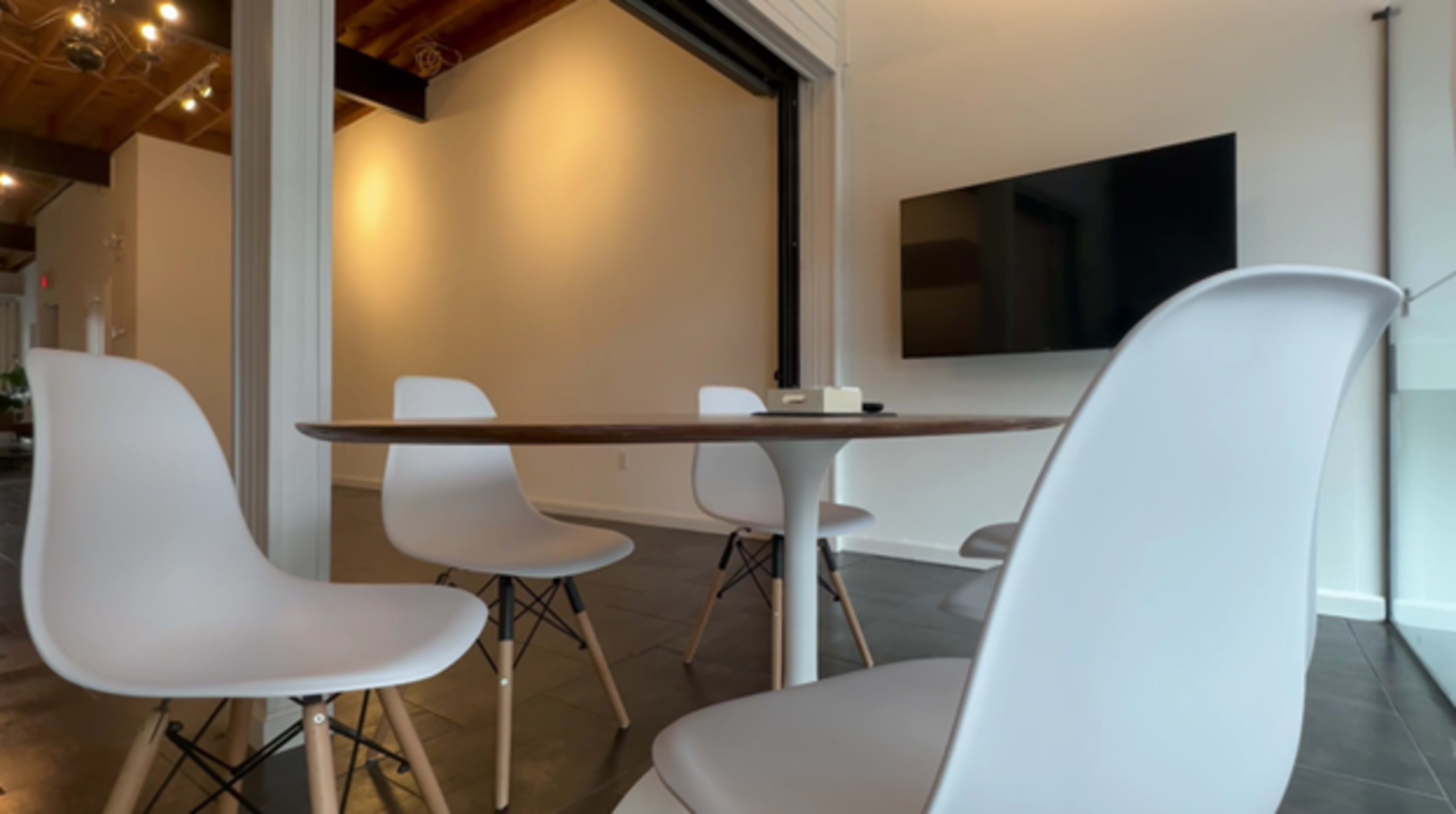 A round table with four white chairs is positioned in a bright, minimalistic room featuring a wall-mounted television.