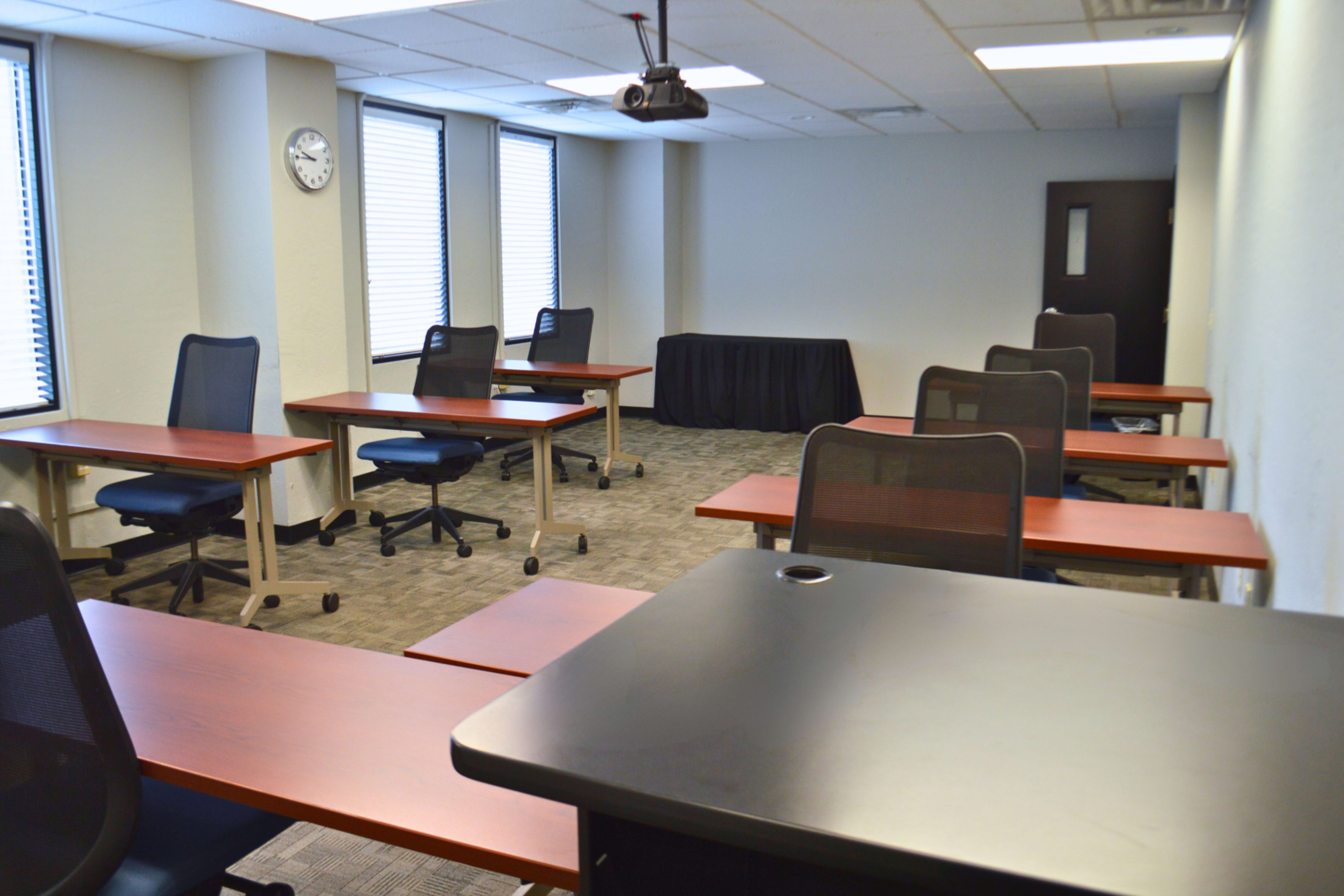A classroom setting with several desks, chairs, and a projector mounted on the ceiling.