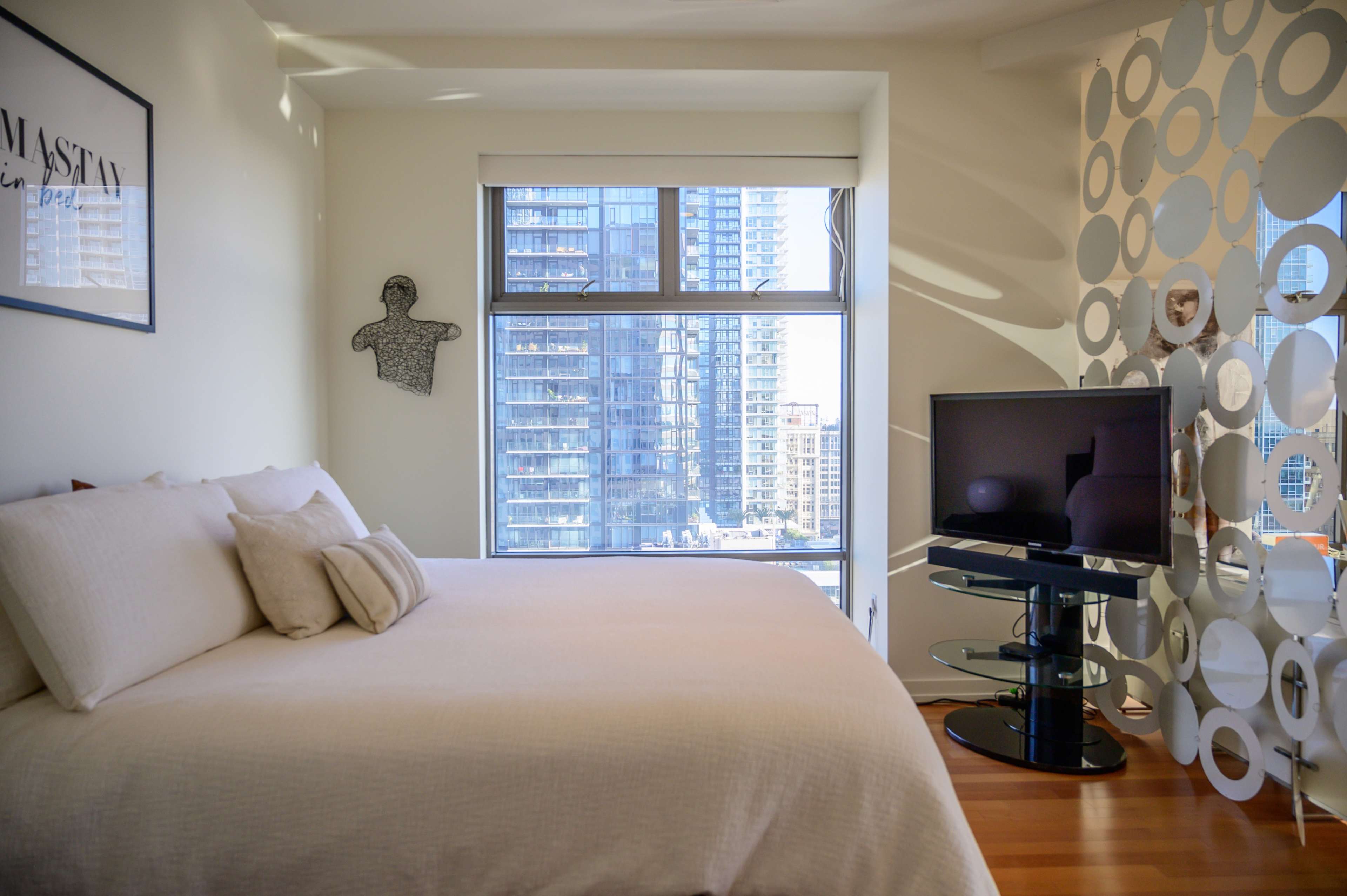 A bedroom with a large bed, a modern television on a glass stand, and a wall decorated with circular patterns, all set in a room with large windows revealing a cityscape.