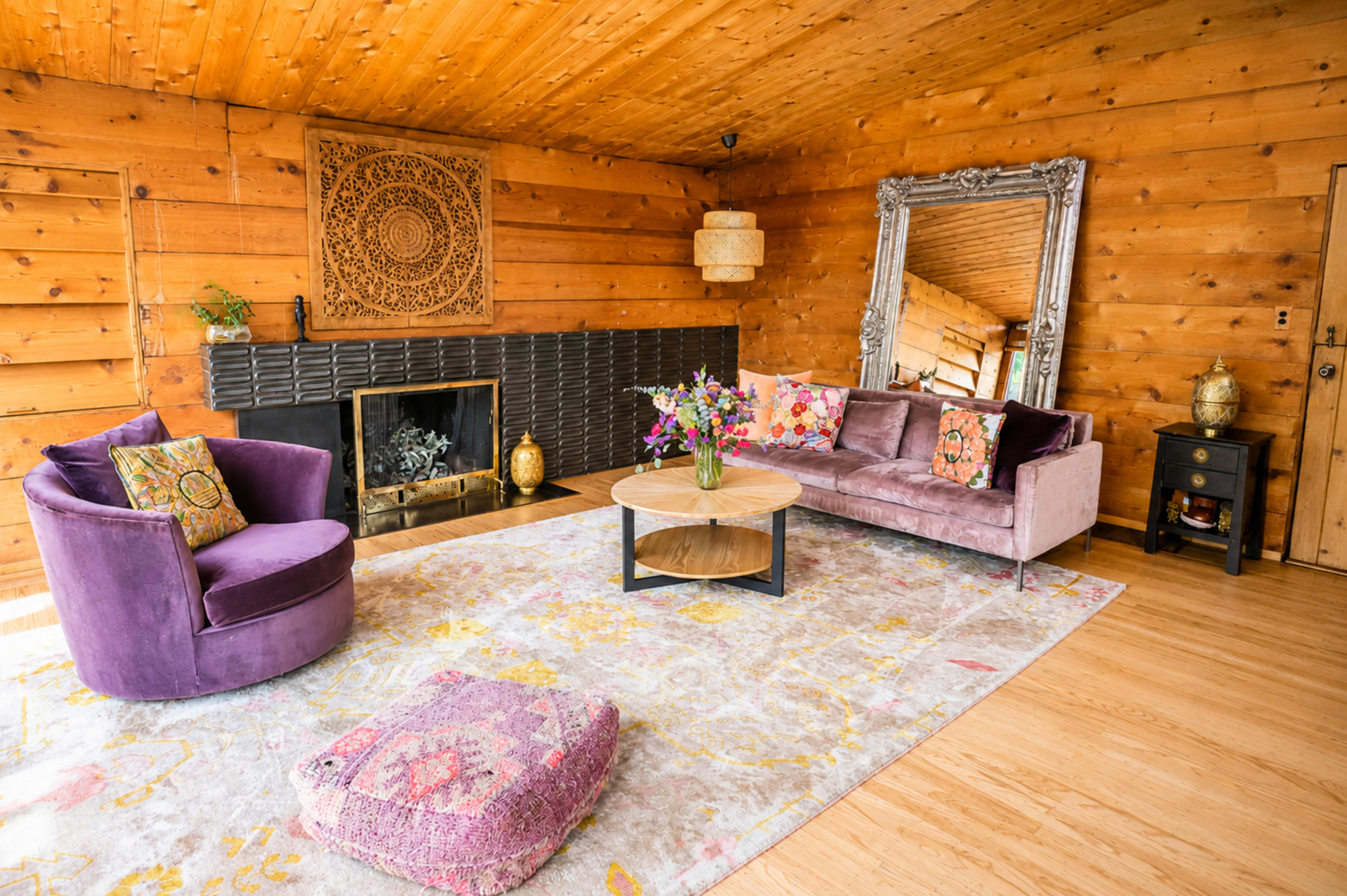 The image displays a cozy living room featuring a purple velvet sofa, a round coffee table, and a decorative fireplace, all set against warm wooden walls.