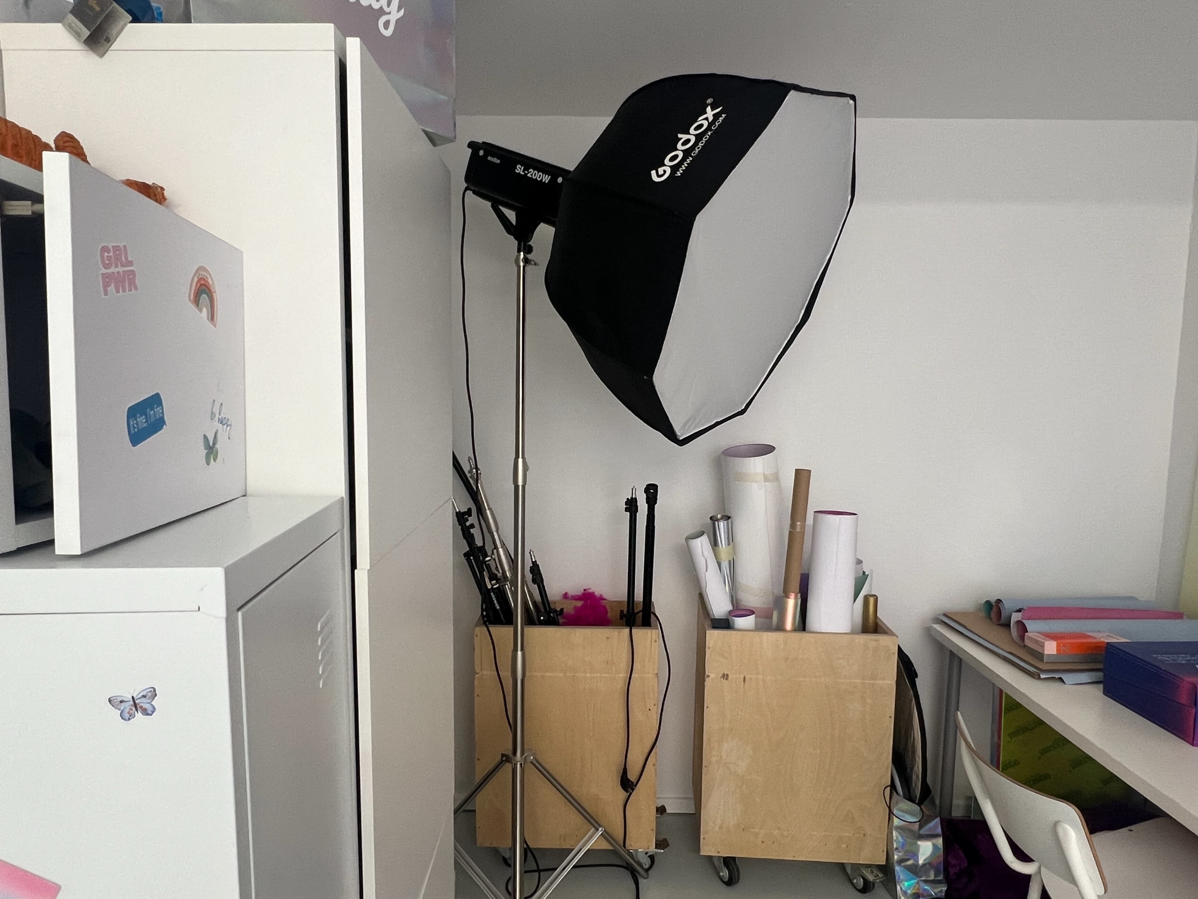 A photography light is positioned next to two wooden storage boxes filled with rolled materials, against a wall in a cluttered room.