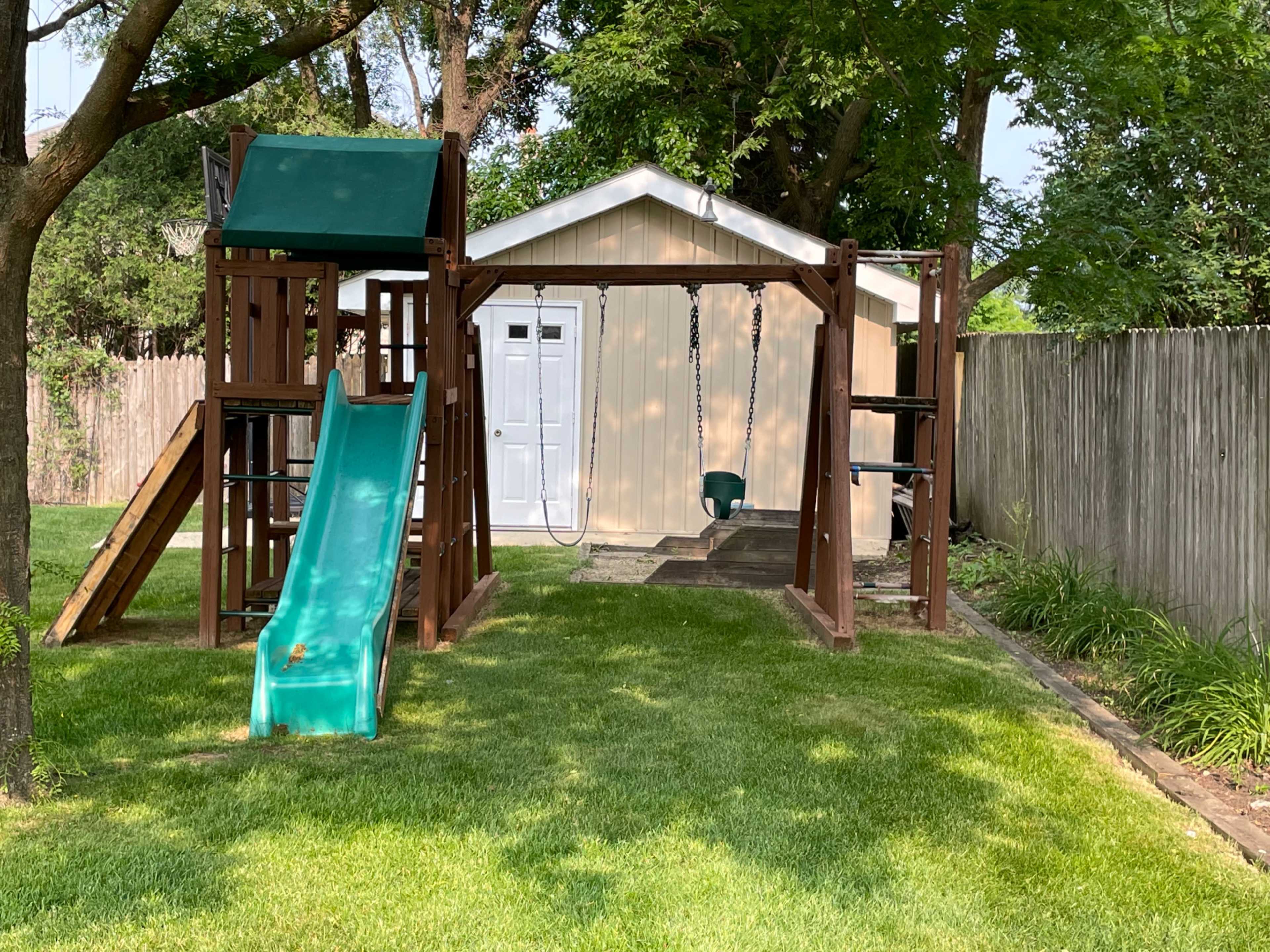 A backyard playset featuring a slide, swings, and a small playhouse, situated on a lawn with green grass.