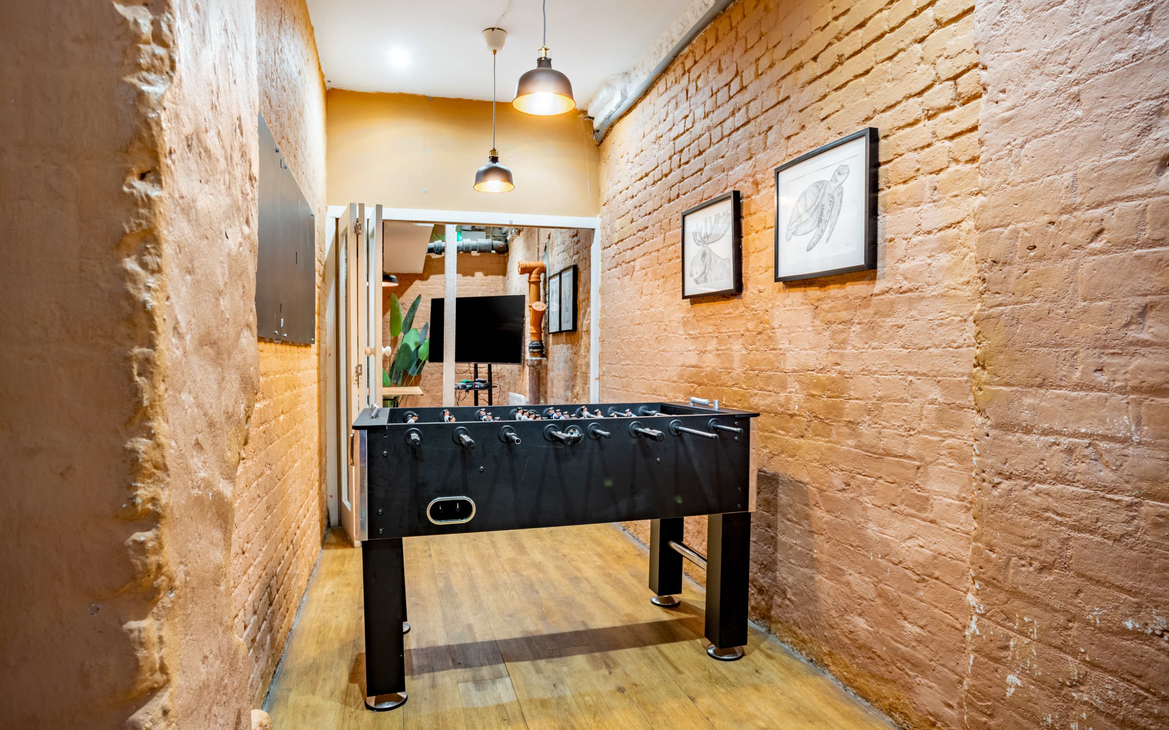 A foosball table is placed in a narrow hallway lined with brick walls and illuminated by overhead lights.