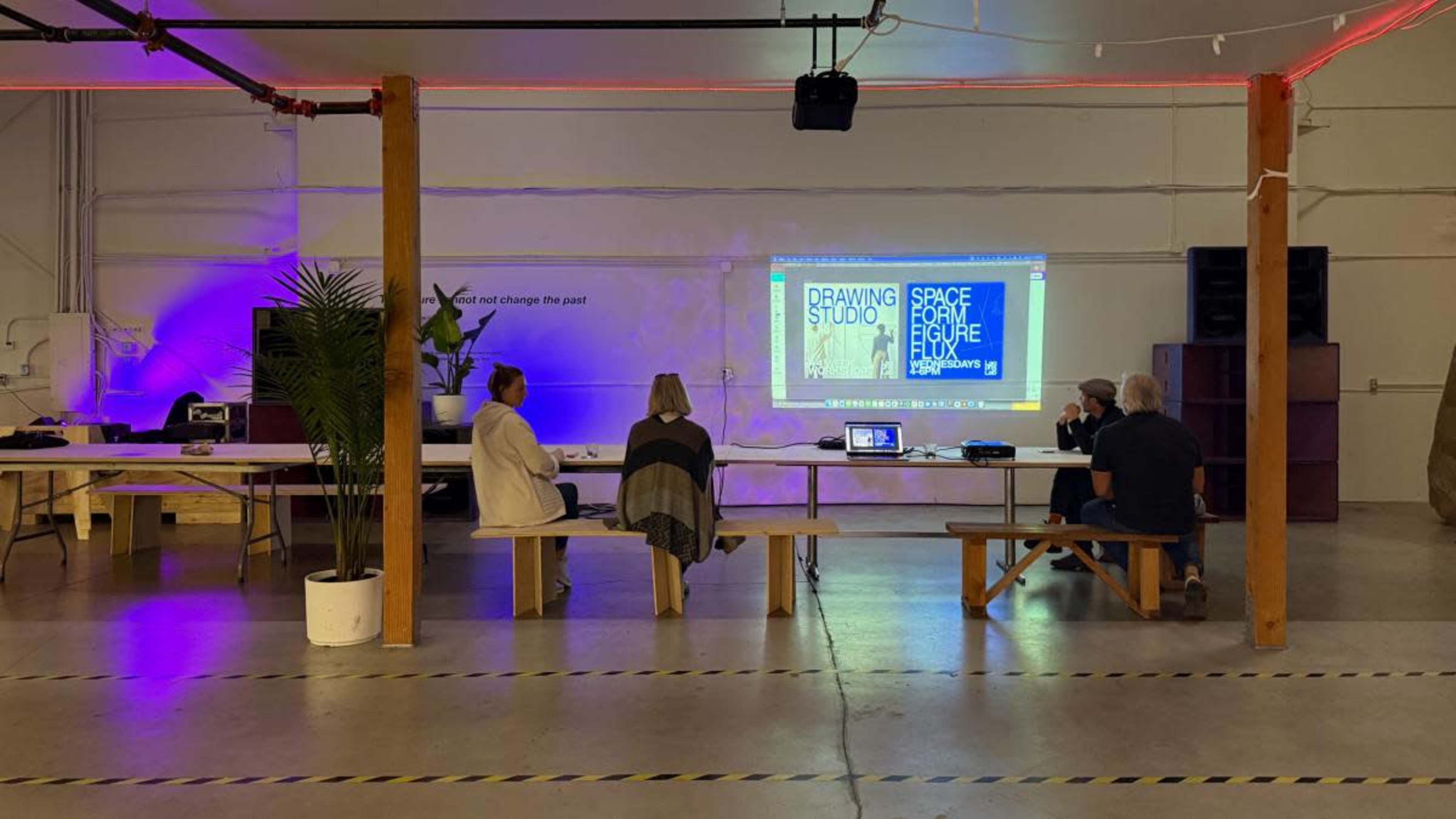 Three people are seated at a long table in a brightly lit space, facing a large screen displaying text about a drawing studio and related topics.