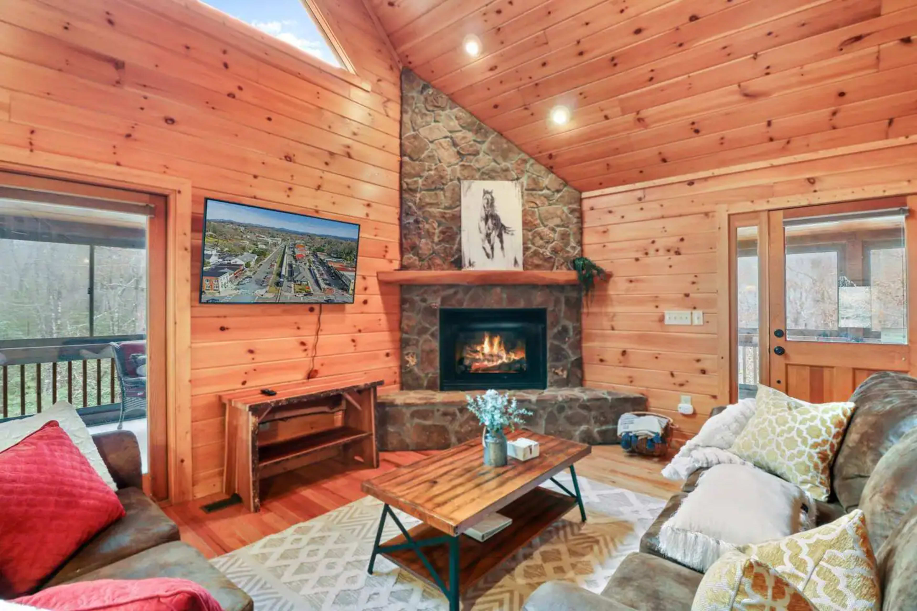 The cozy living room features wooden walls, a stone fireplace with a fire burning, and a television mounted above a rustic coffee table.