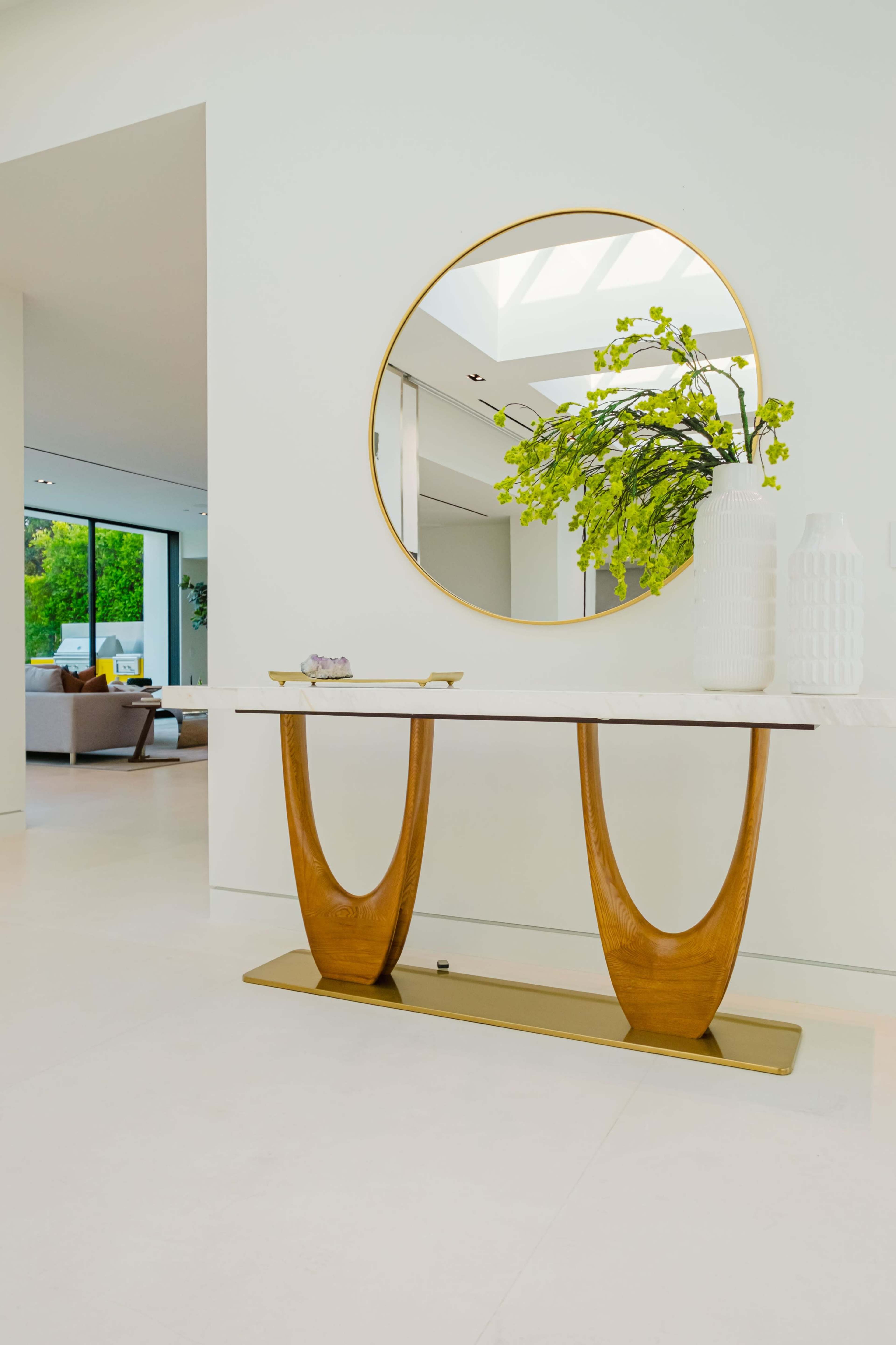A modern console table with a marble top and wooden base stands against a white wall, complemented by a round mirror and vases containing greenery.