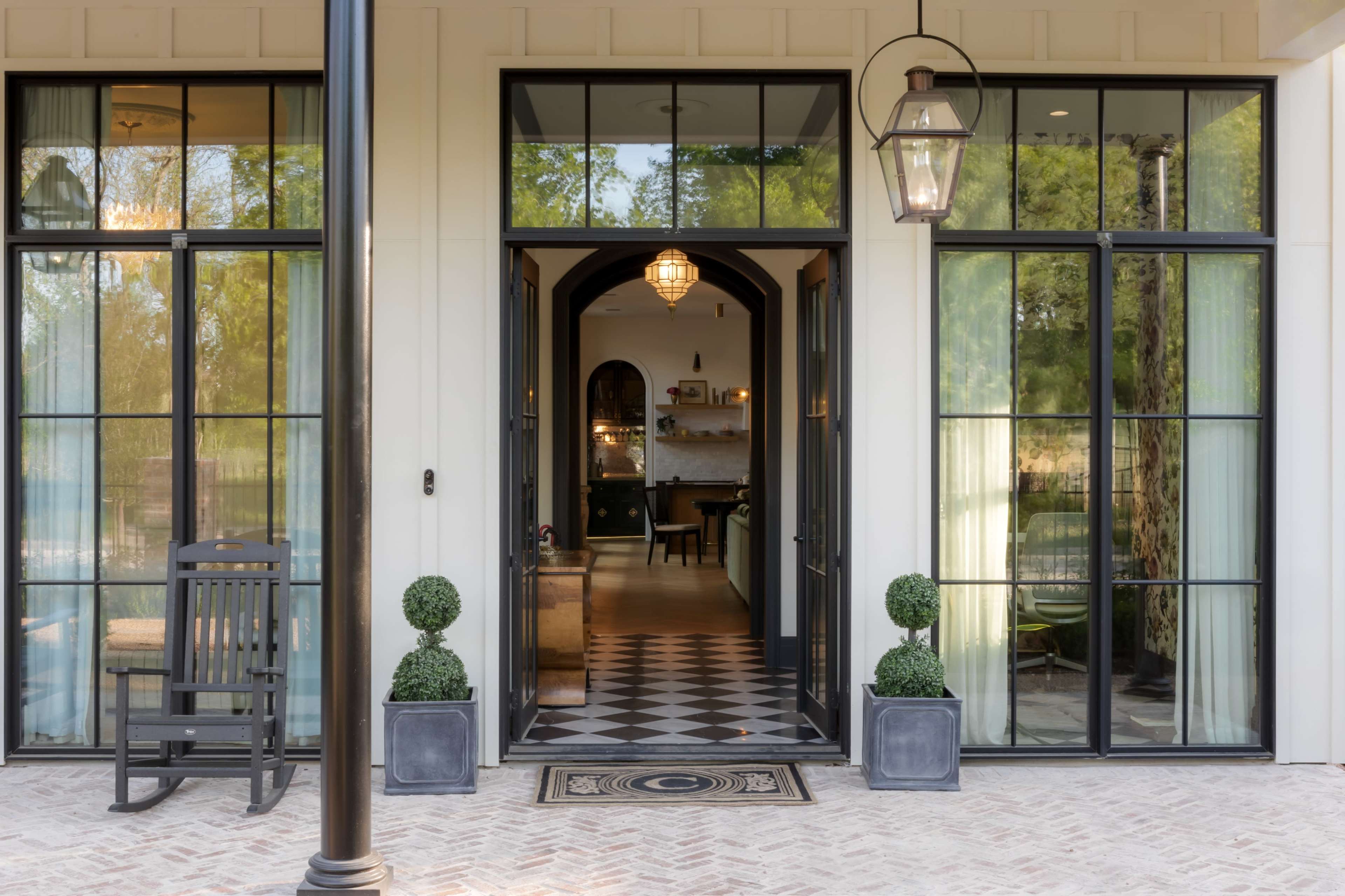 The image shows the entrance of a house with glass doors, decorative planters, and a checkered floor leading into a well-lit interior.