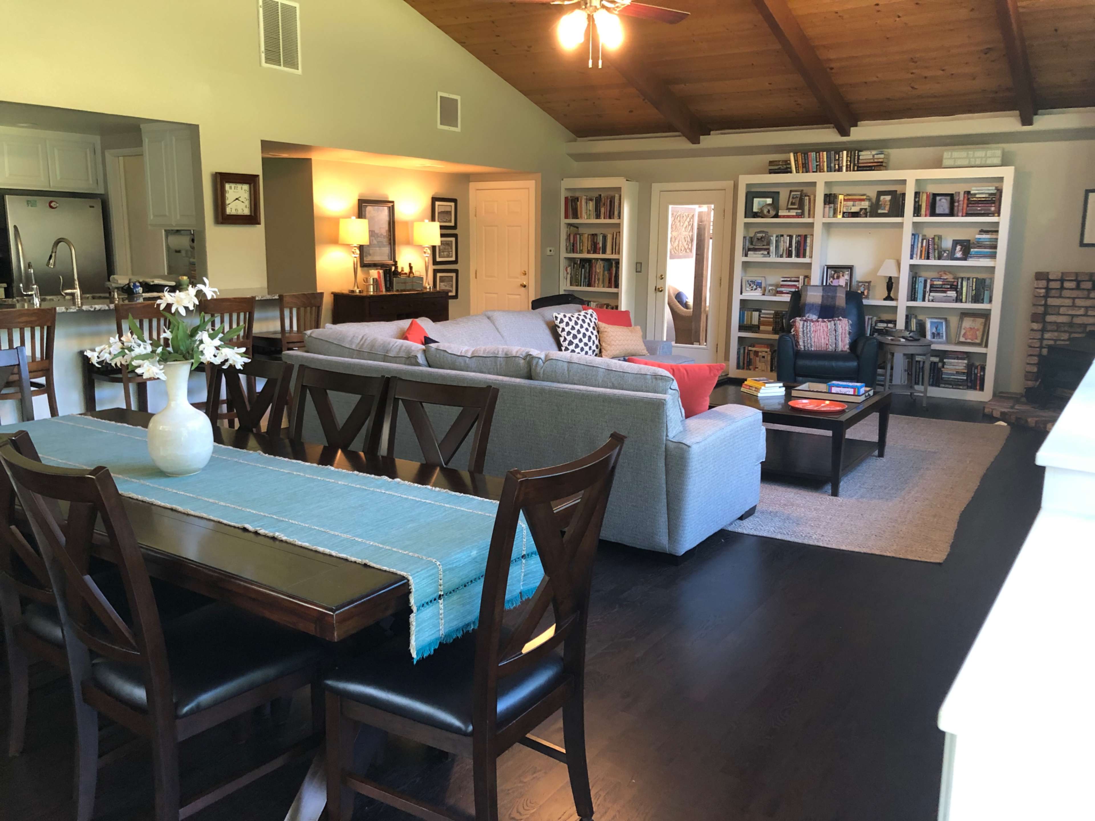 A spacious living area featuring a gray sectional sofa, a coffee table, bookshelves filled with books, and a dining table with chairs, all under a wooden ceiling.