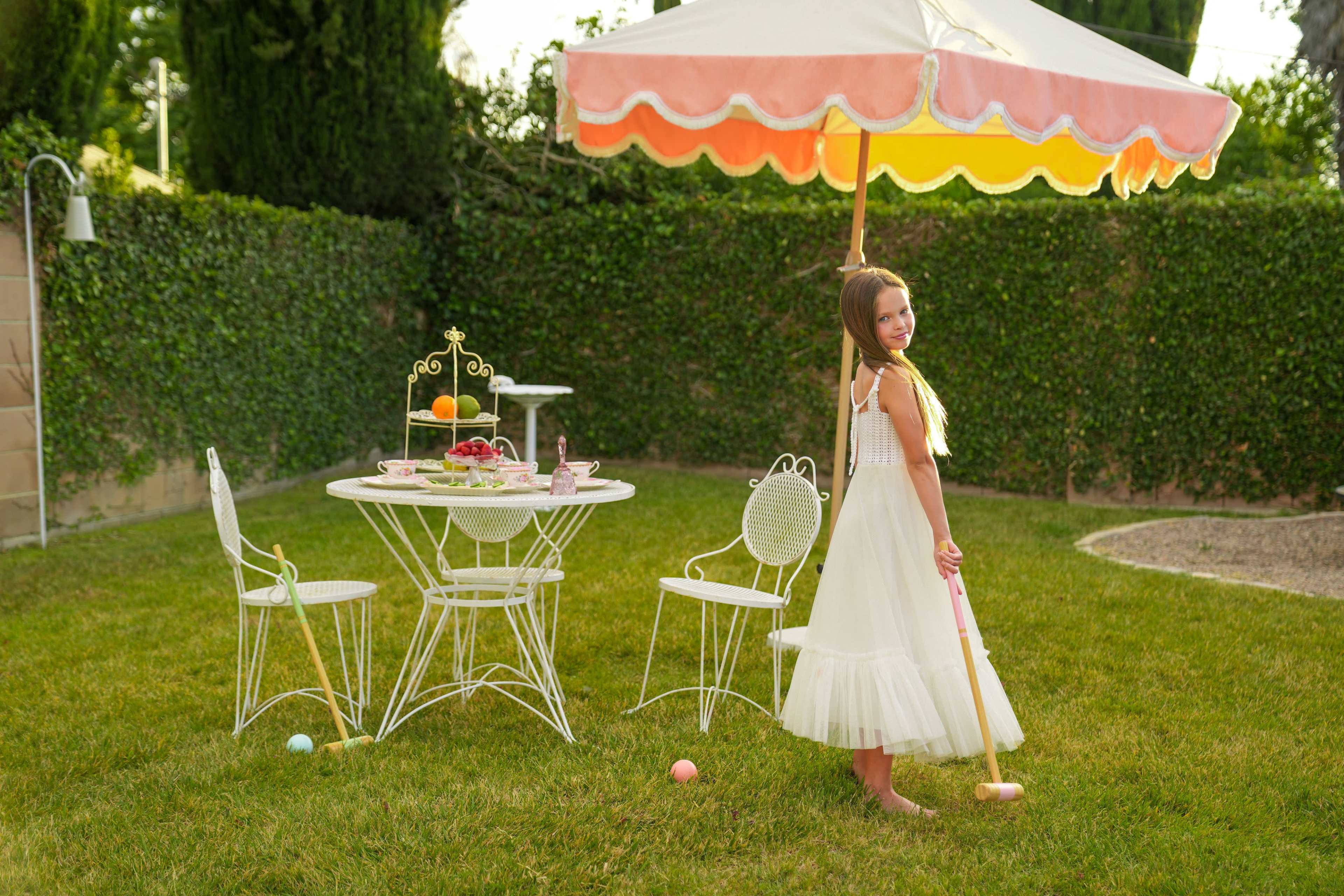 A young girl in a white dress stands with a croquet mallet beside an outdoor table set with refreshments and surrounded by green grass and a large umbrella.