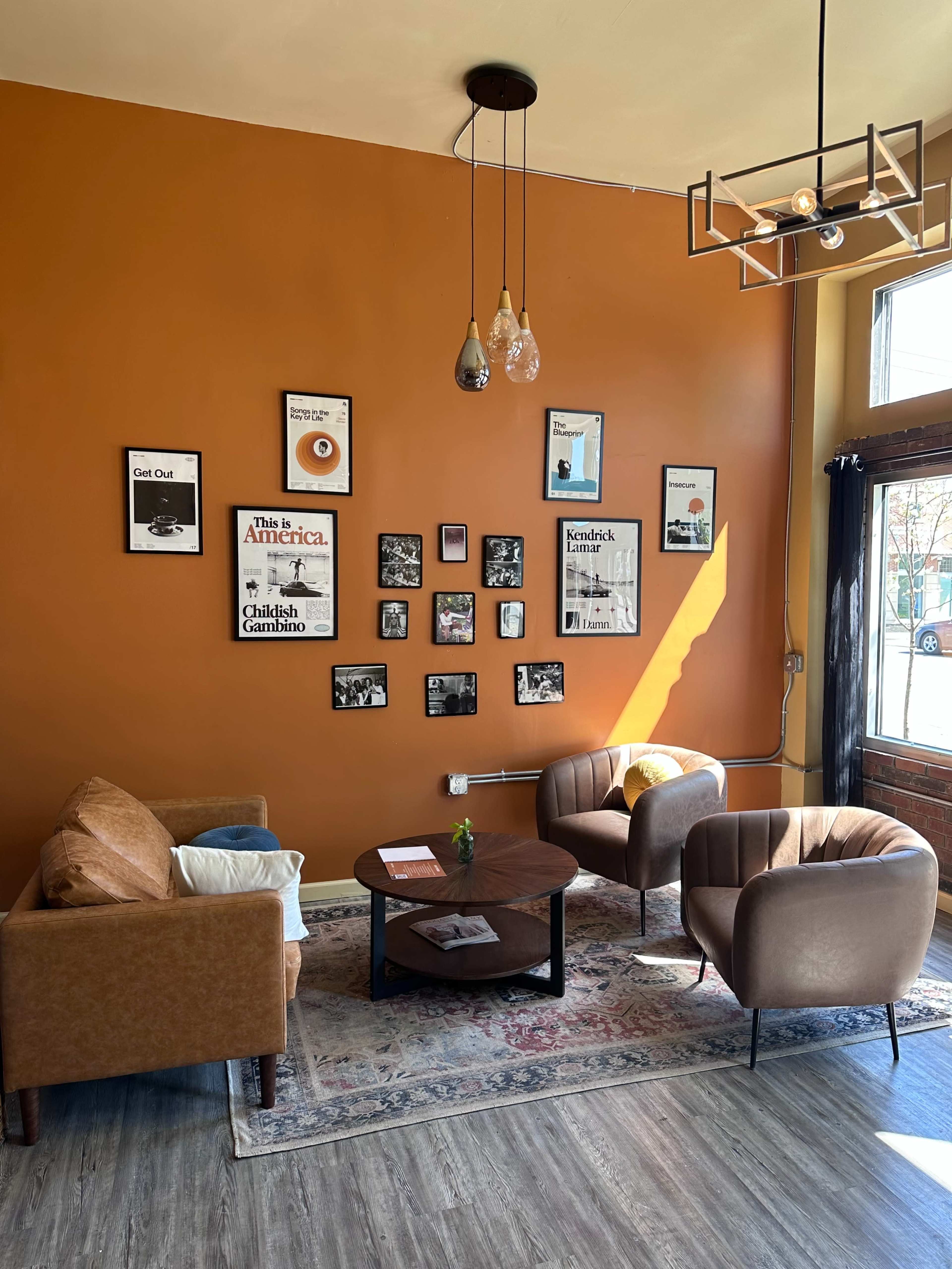 A cozy seating area features two brown armchairs and a small round table, against an orange wall adorned with framed posters and photographs.