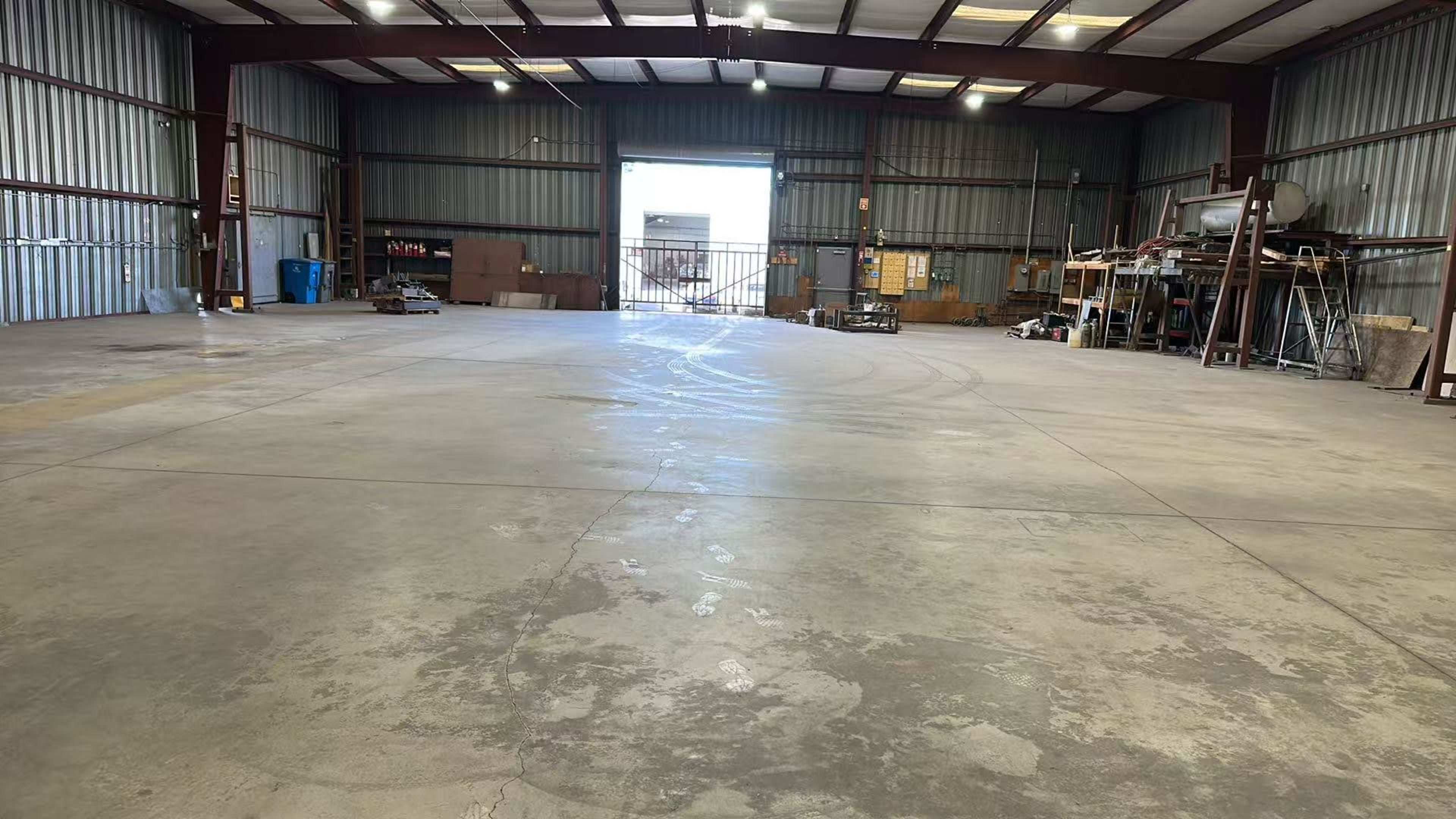 The image shows a spacious industrial warehouse with metal walls, a high ceiling, and a wide open area leading to a large entrance.