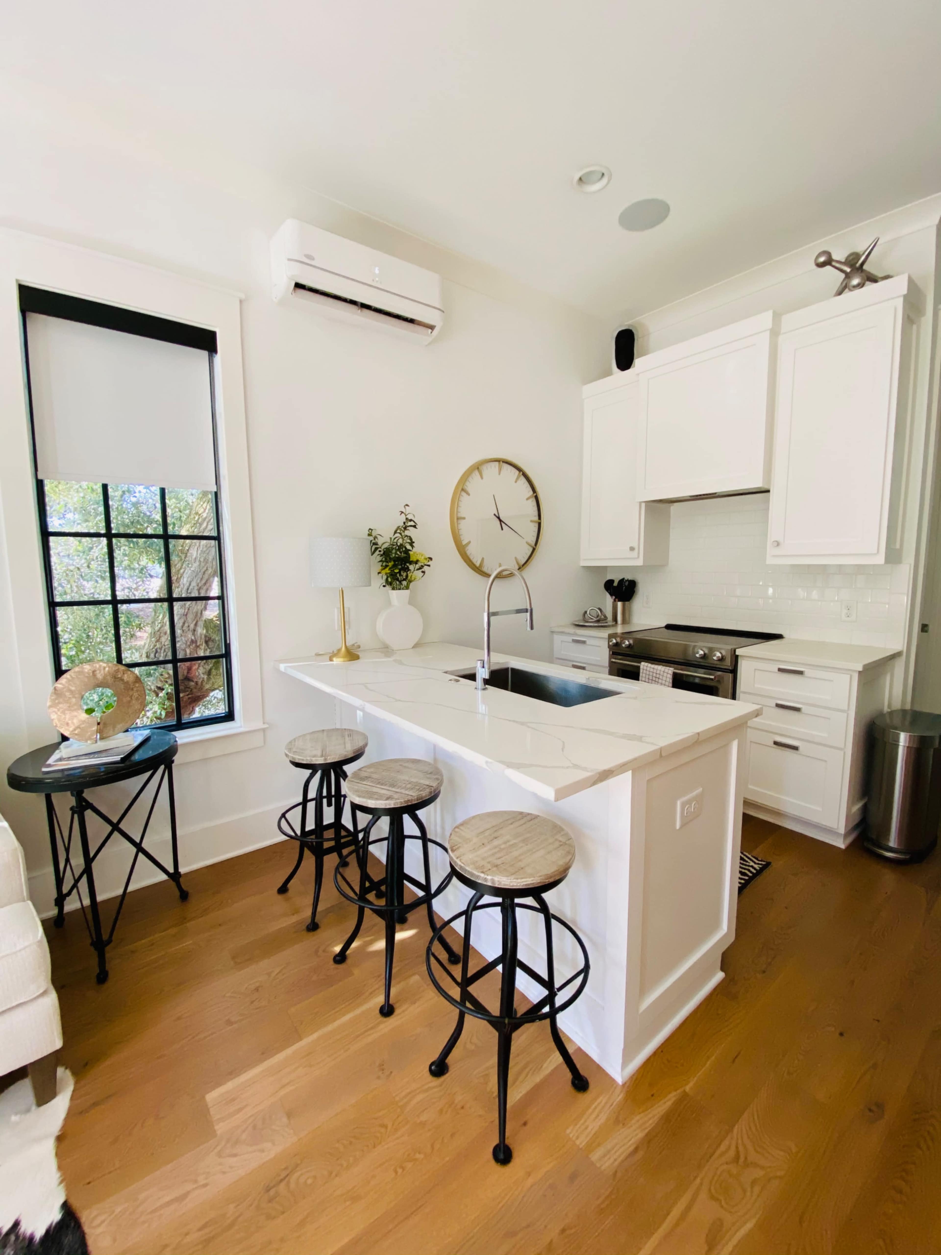A modern kitchen features a white island with three wooden bar stools, stainless steel appliances, and a clock on the wall.