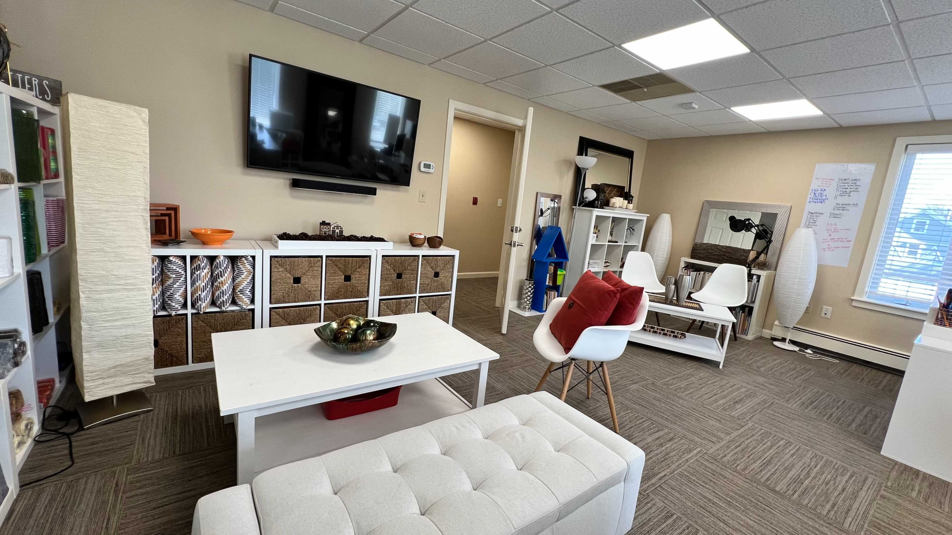 A modern, organized office space featuring a TV mounted on the wall, a white coffee table, and various seating options with light-colored furniture and decorative elements.