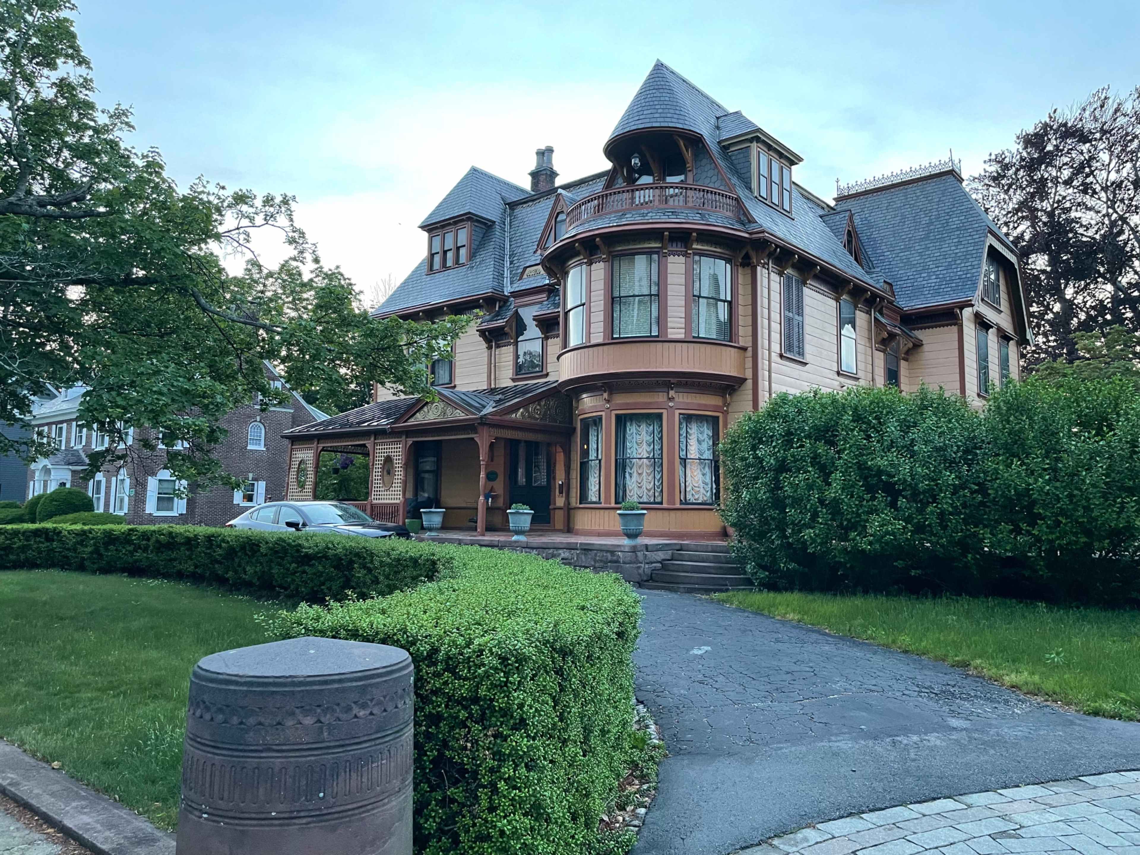 A large Victorian-style house with a rounded turret and intricate woodwork, surrounded by green shrubbery and a manicured lawn.