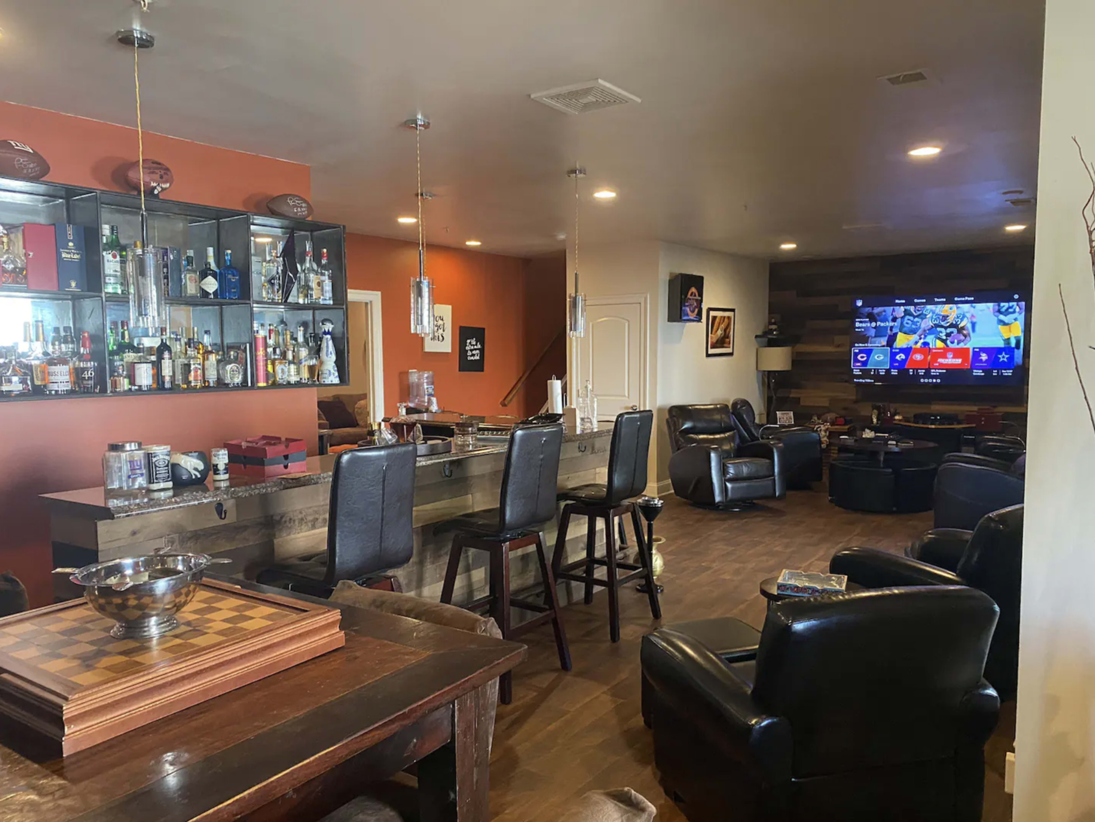 A spacious entertainment area featuring a bar with stools, a large television, leather seating, and shelves displaying various bottles.