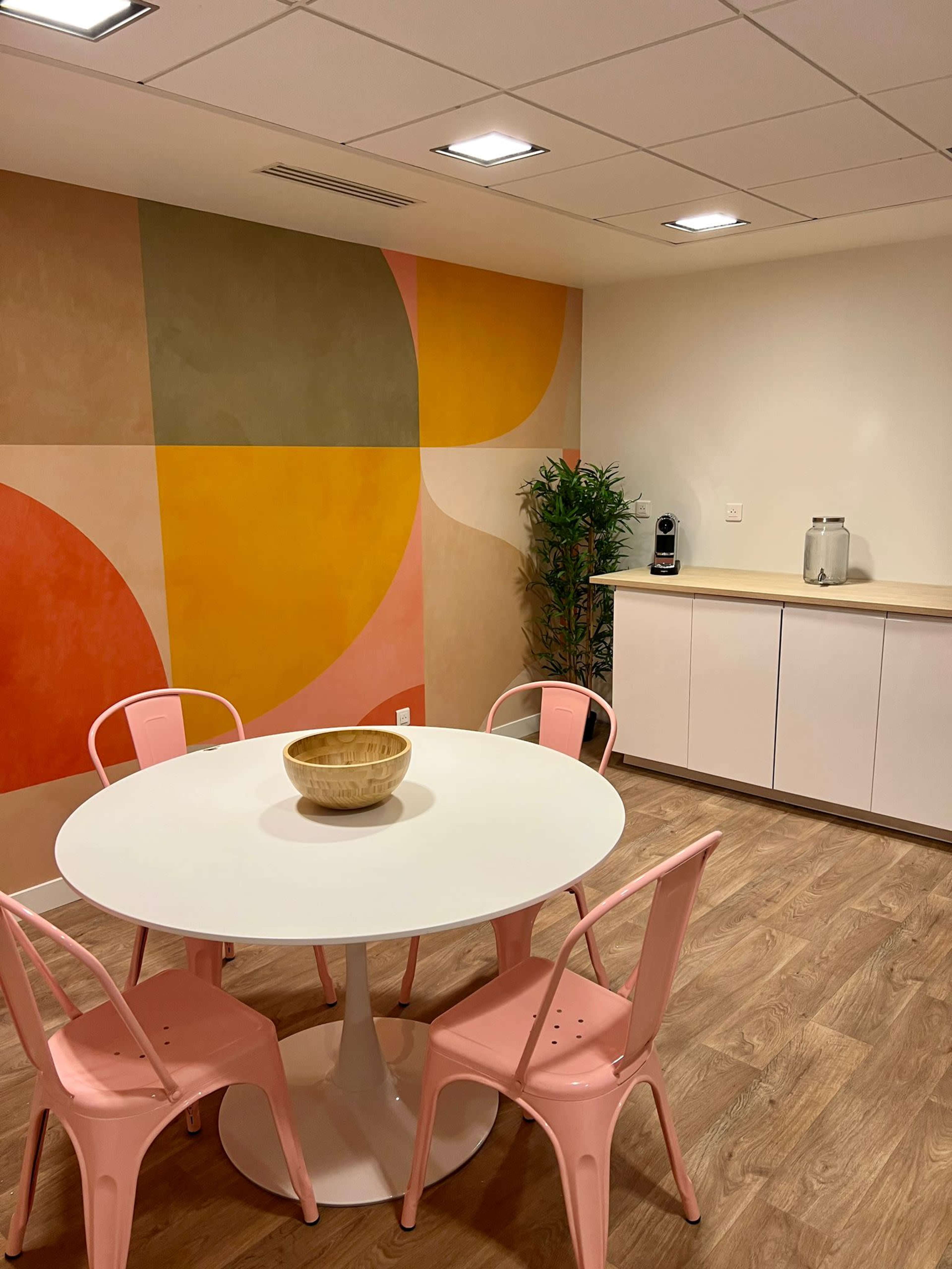 A minimalist dining area with a round white table at the center, surrounded by pink chairs, against a backdrop of colorful geometric wall art.