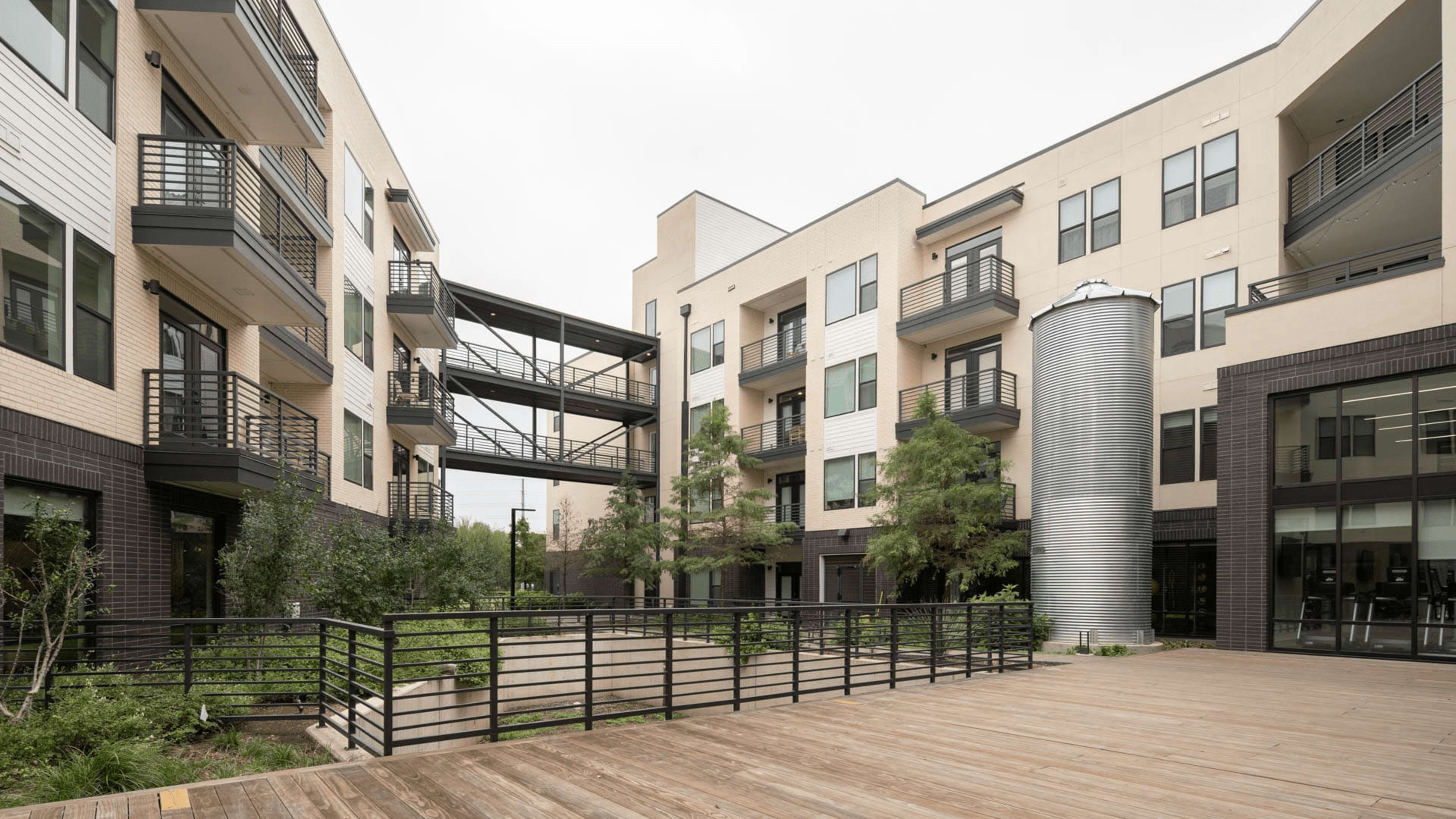 A modern courtyard features multiple apartment buildings with balconies, a metal structure, and wooden decking surrounded by greenery.