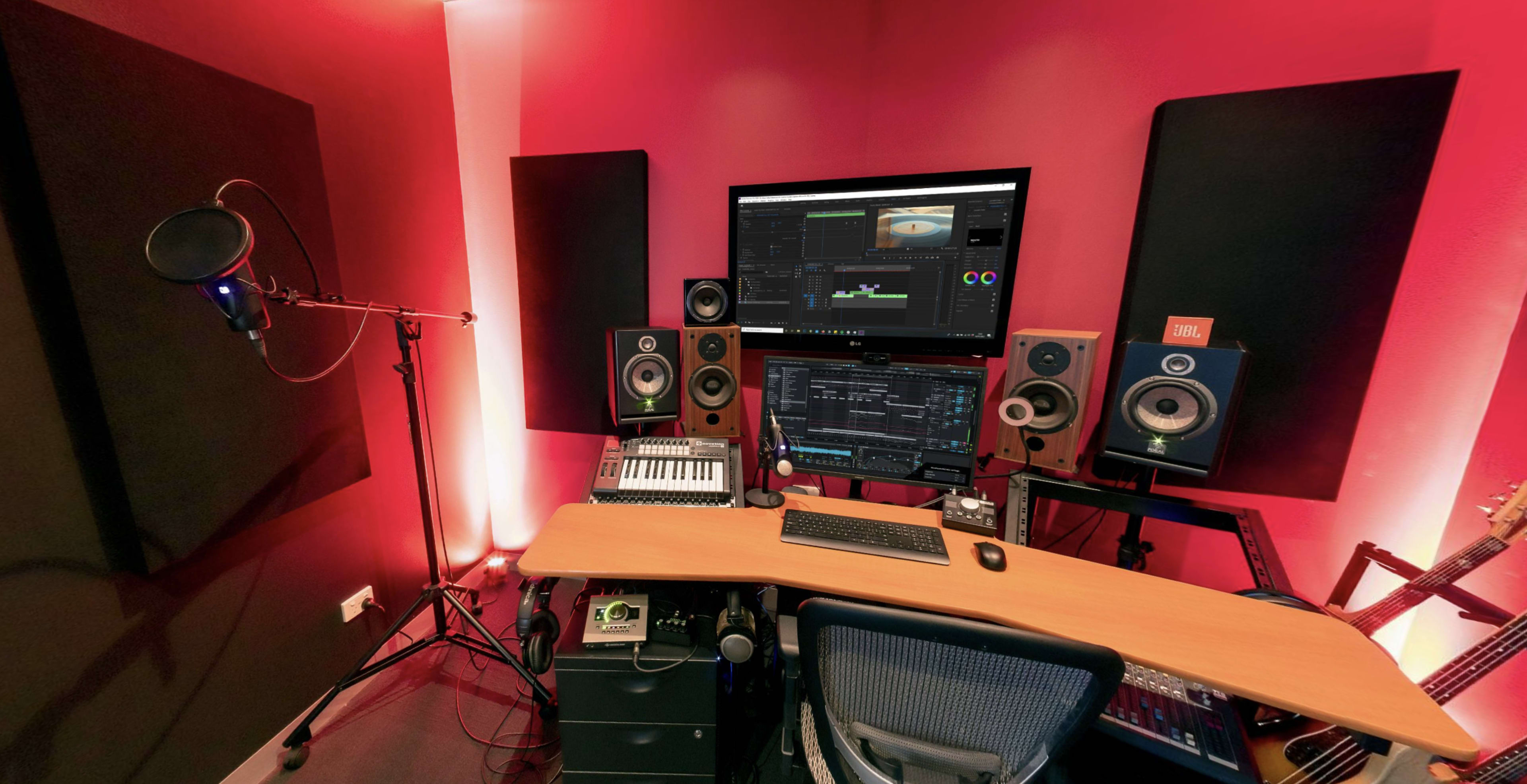 The image shows a music production studio featuring a microphone, computer monitor, speakers, and a keyboard arranged on a wooden desk against red-lit walls.