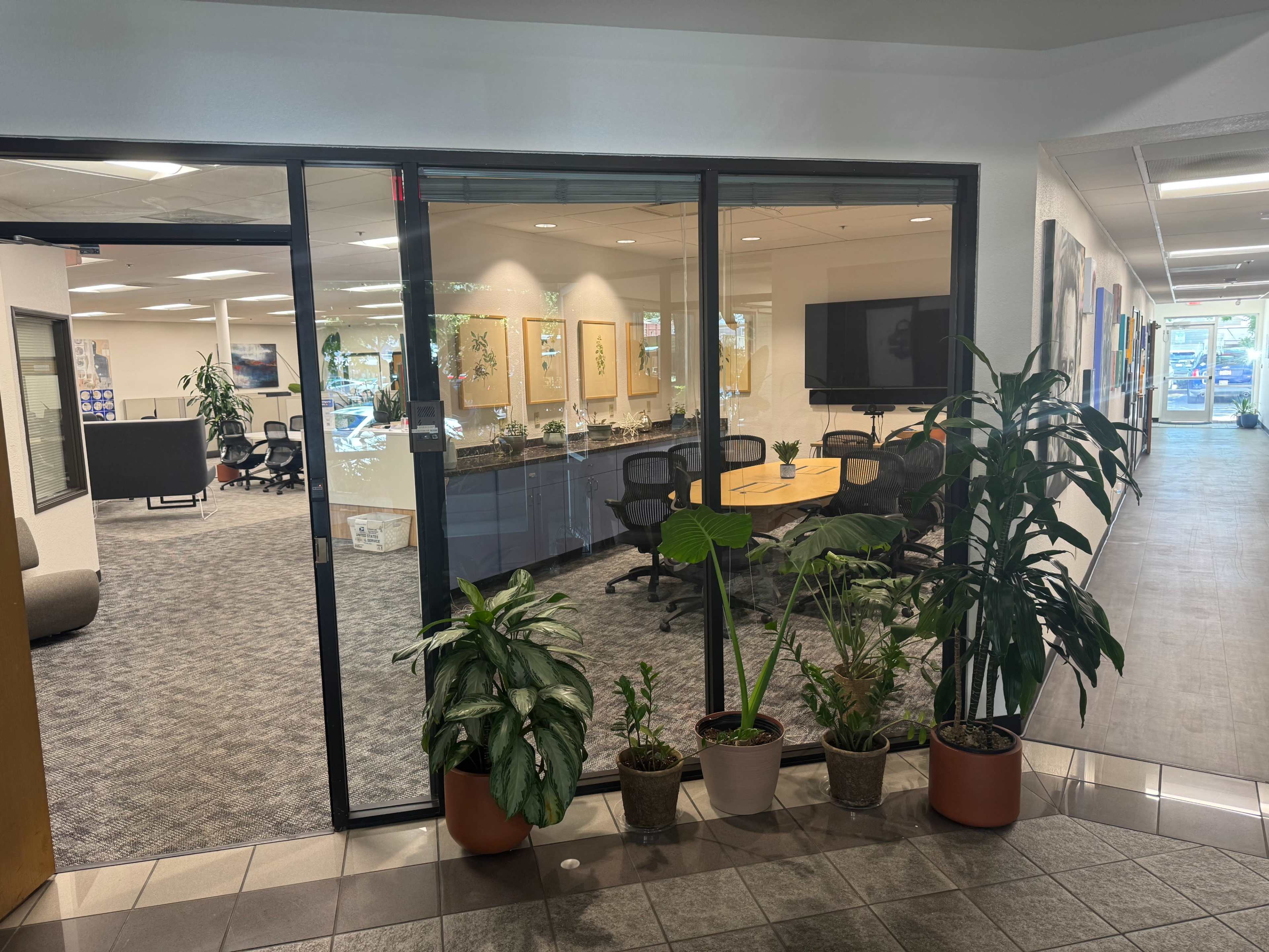 The image shows a modern office space with a conference room visible through a large glass wall, surrounded by potted plants in a corridor.