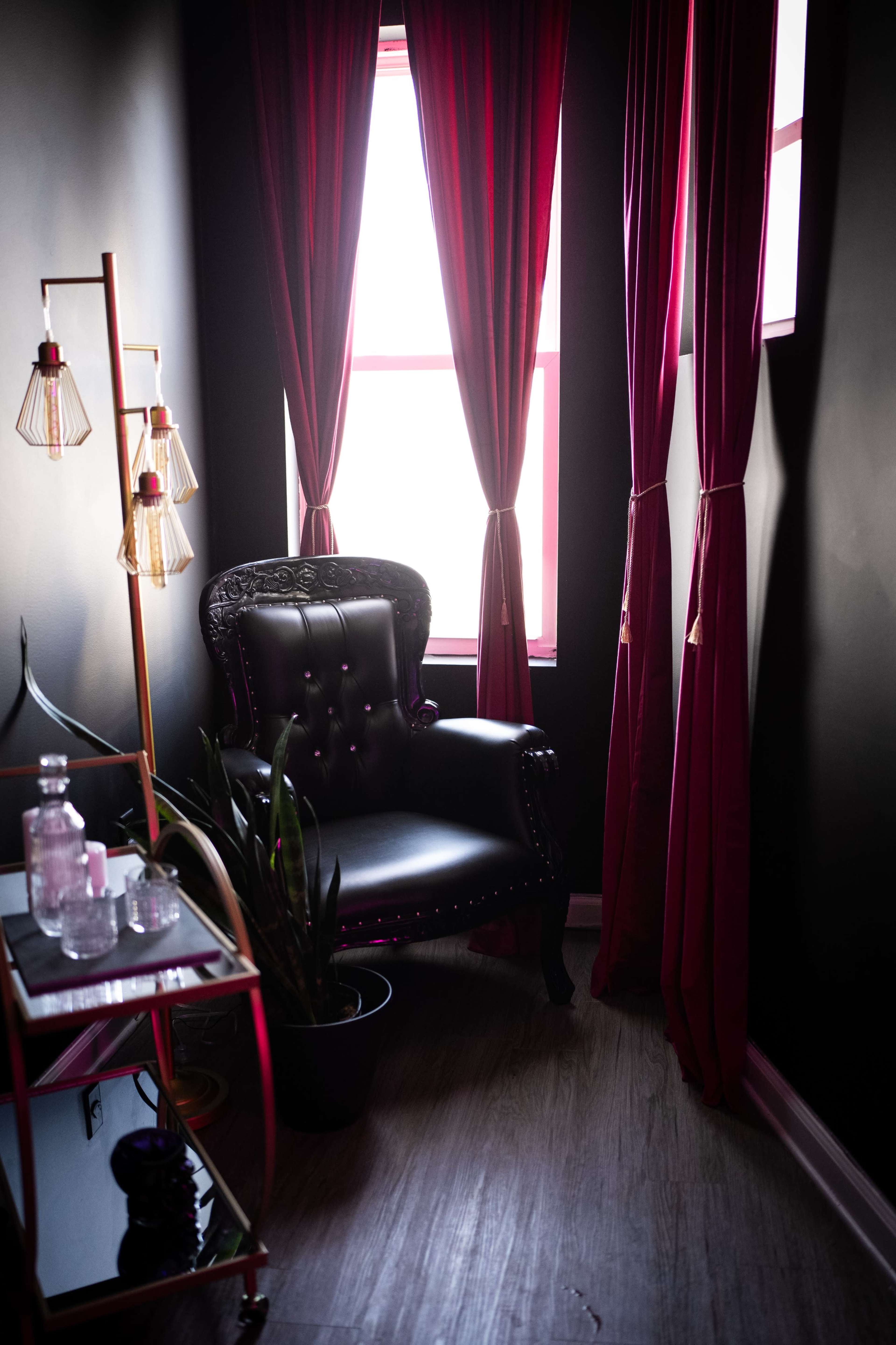 A black upholstered armchair sits beside a plant and a small glass table, framed by large, deep red curtains in a dimly lit corner.