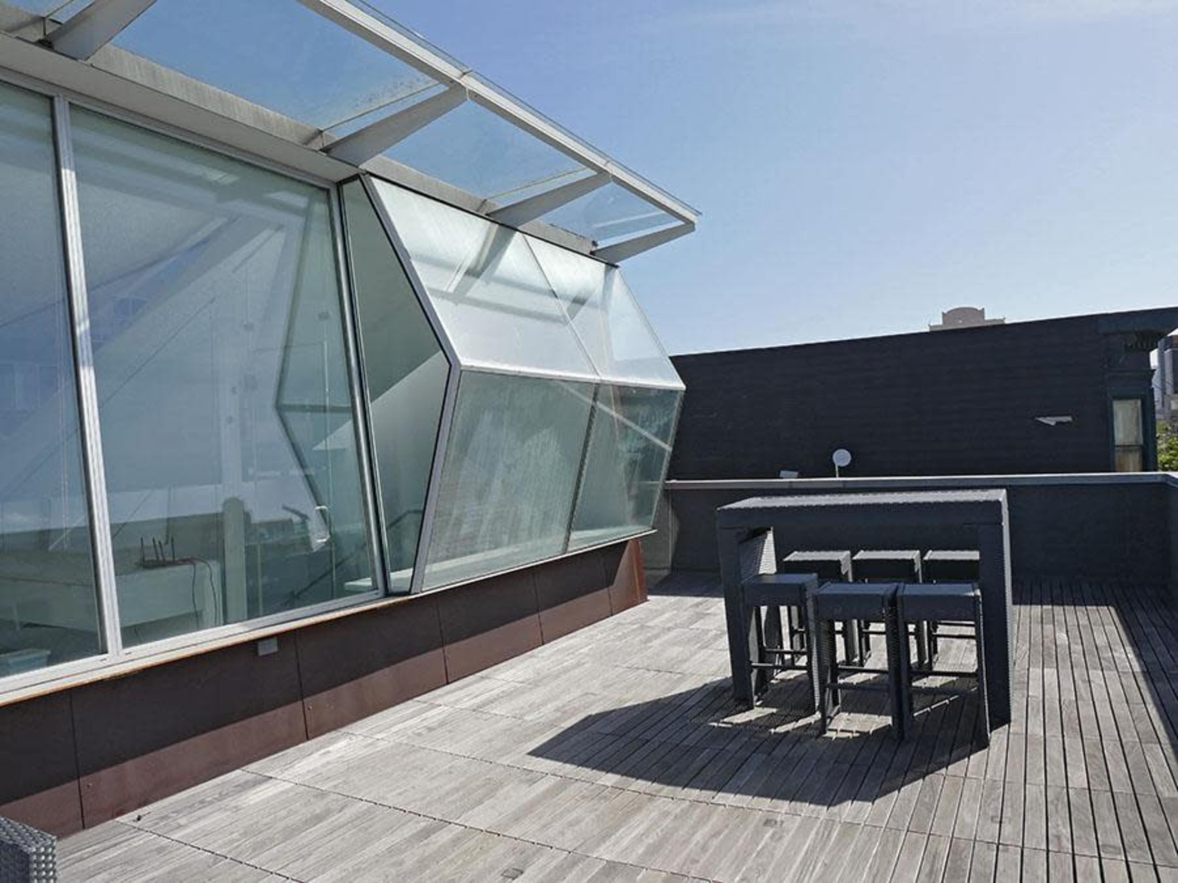 A modern rooftop terrace featuring a glass-walled structure and an outdoor dining area with a table and chairs.