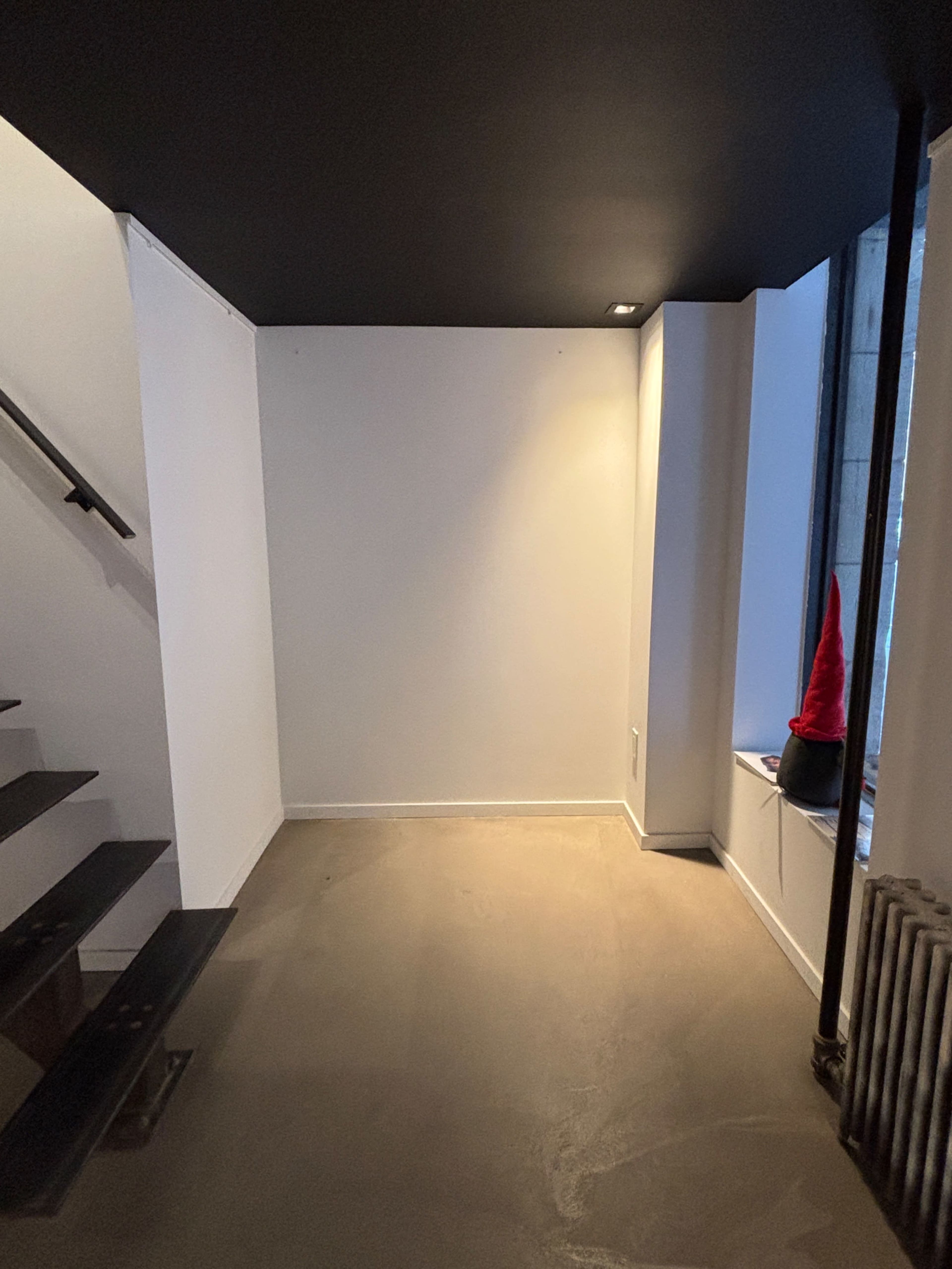 The image shows an empty room with a black ceiling, white walls, a staircase on the left, and a window with a red object in the corner.
