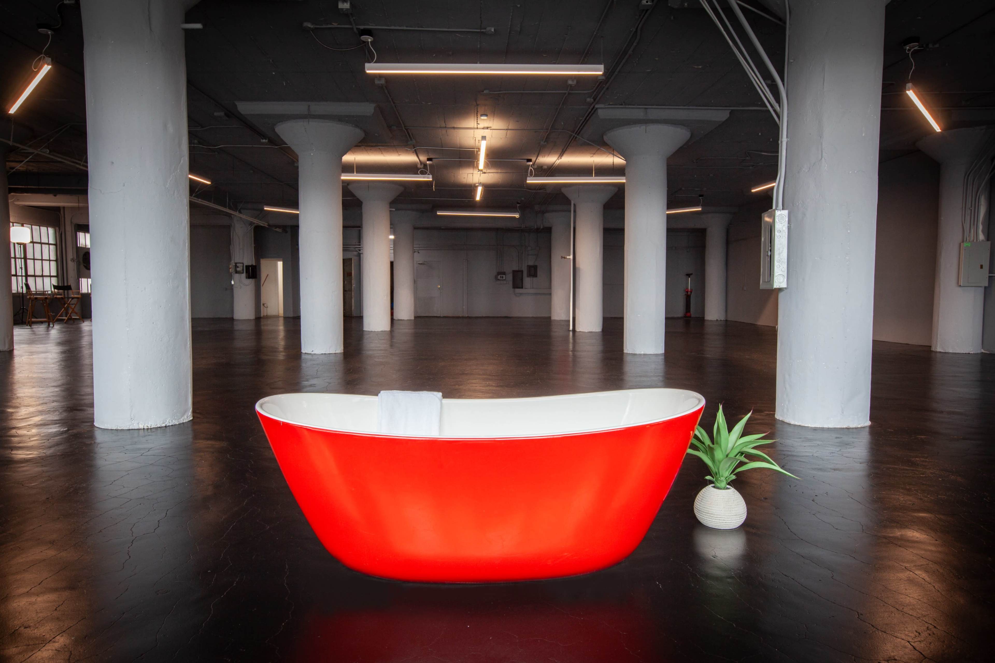 A bright red freestanding bathtub is positioned in the center of a spacious, empty industrial-style room with concrete pillars and polished black floors.