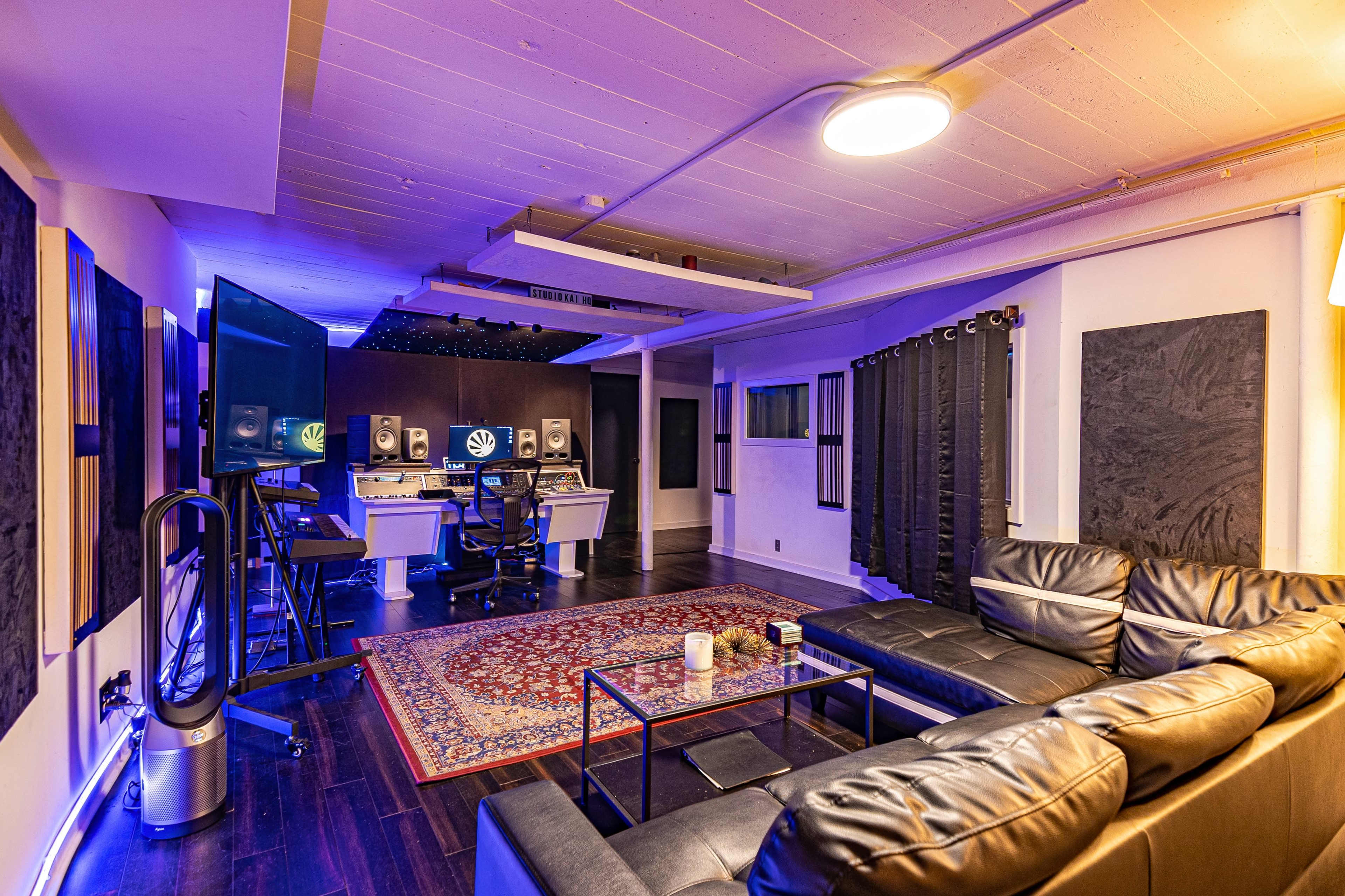 A well-equipped music studio featuring two computer stations, black acoustic panels, a seating area with a couch, and ambient lighting.