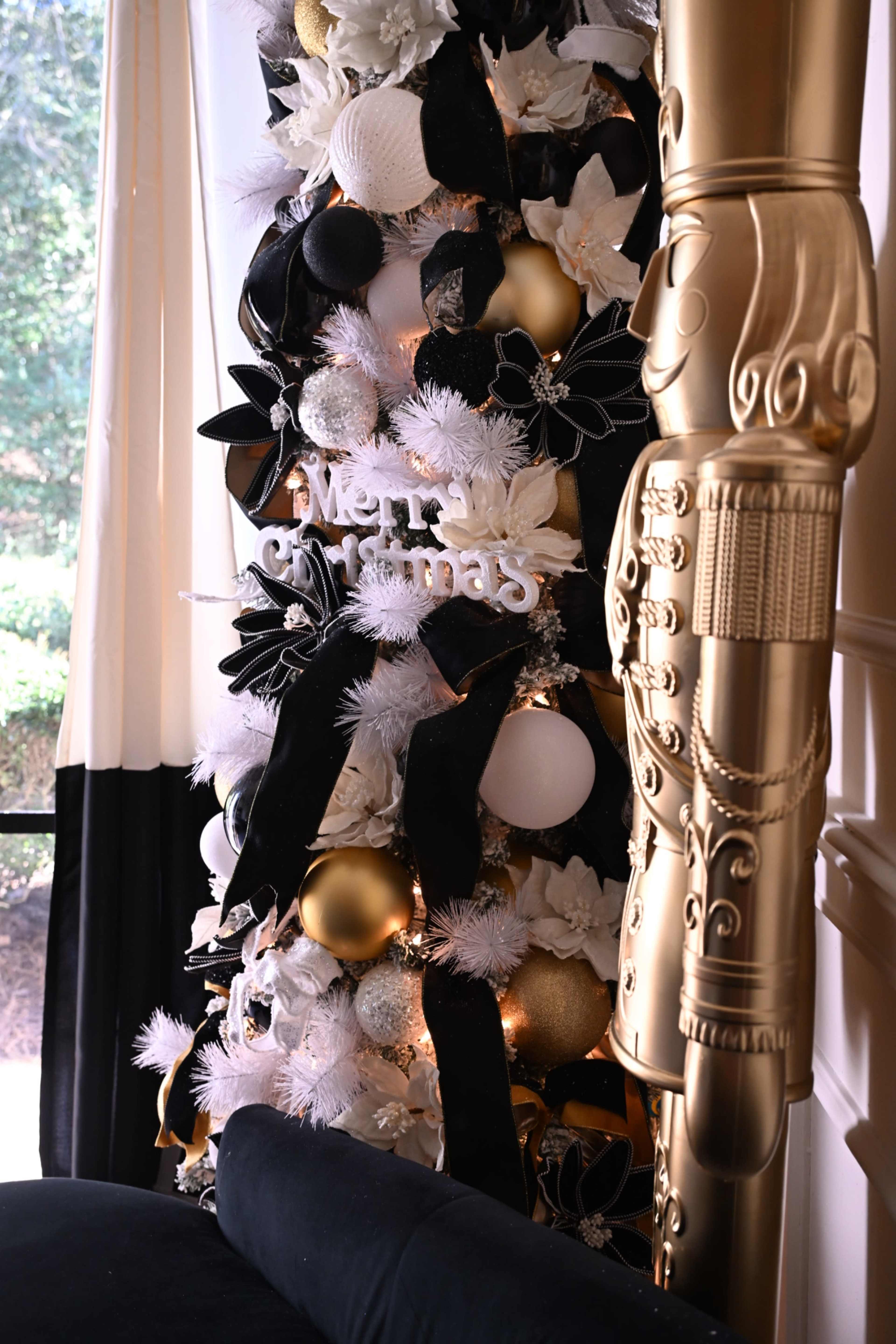 A decorated Christmas tree with black and white ornaments and a "Merry Christmas" sign stands beside a golden nutcracker piece.