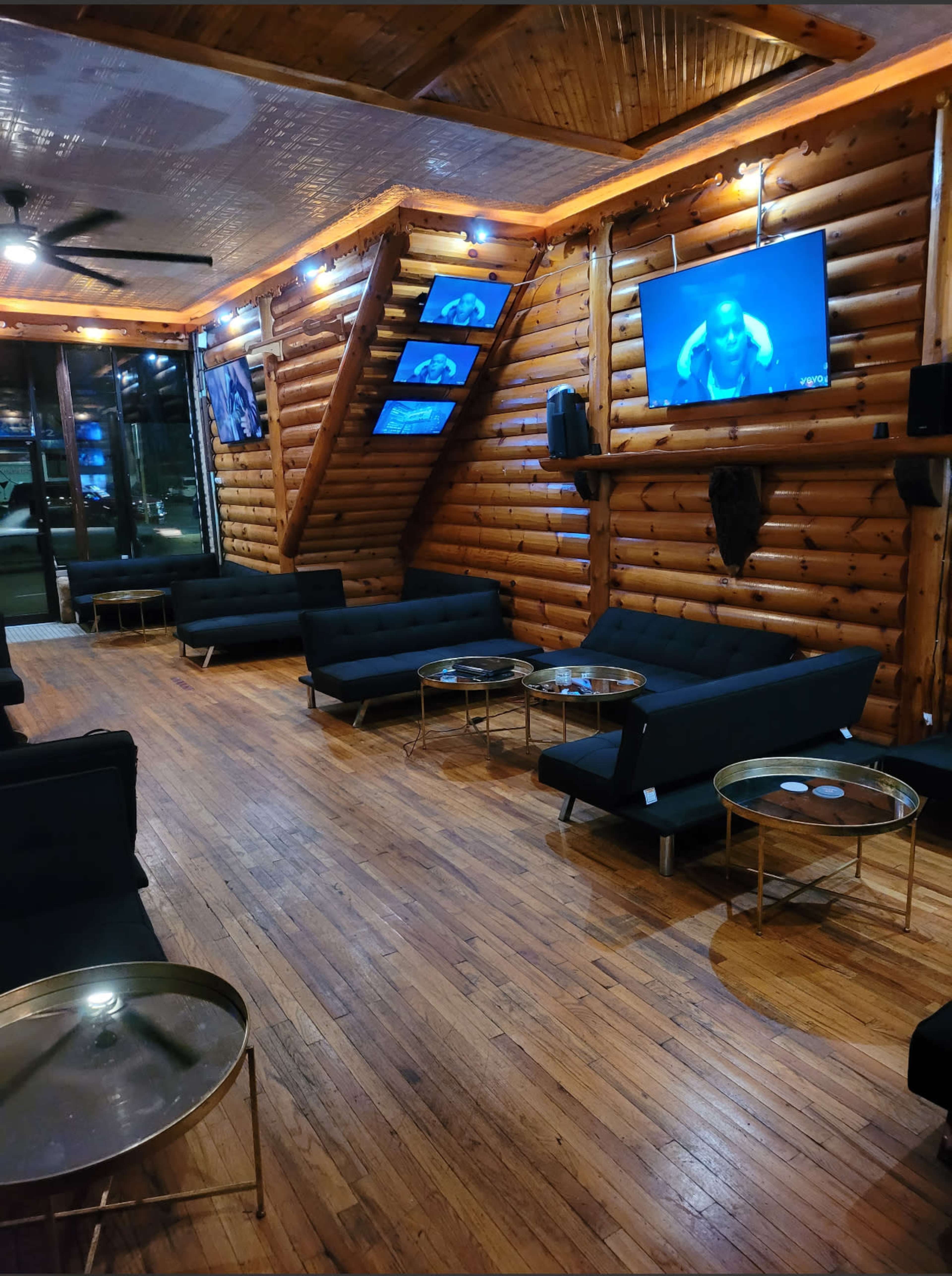A wooden interior lounge area with black sofas, low glass tables, and multiple screens displaying content on the walls.