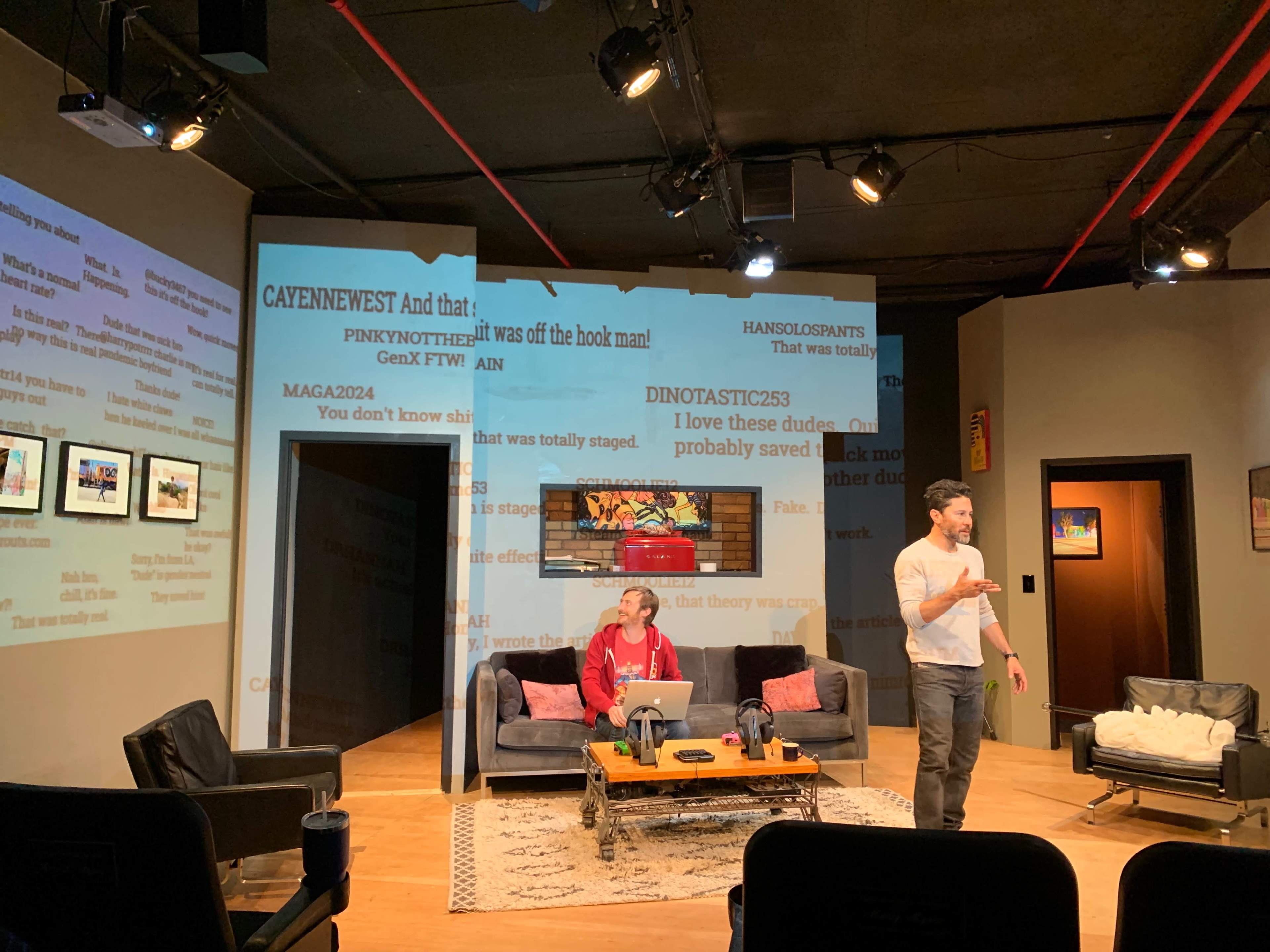 A man stands on stage while a woman sits on a couch, with large text projected on the background.