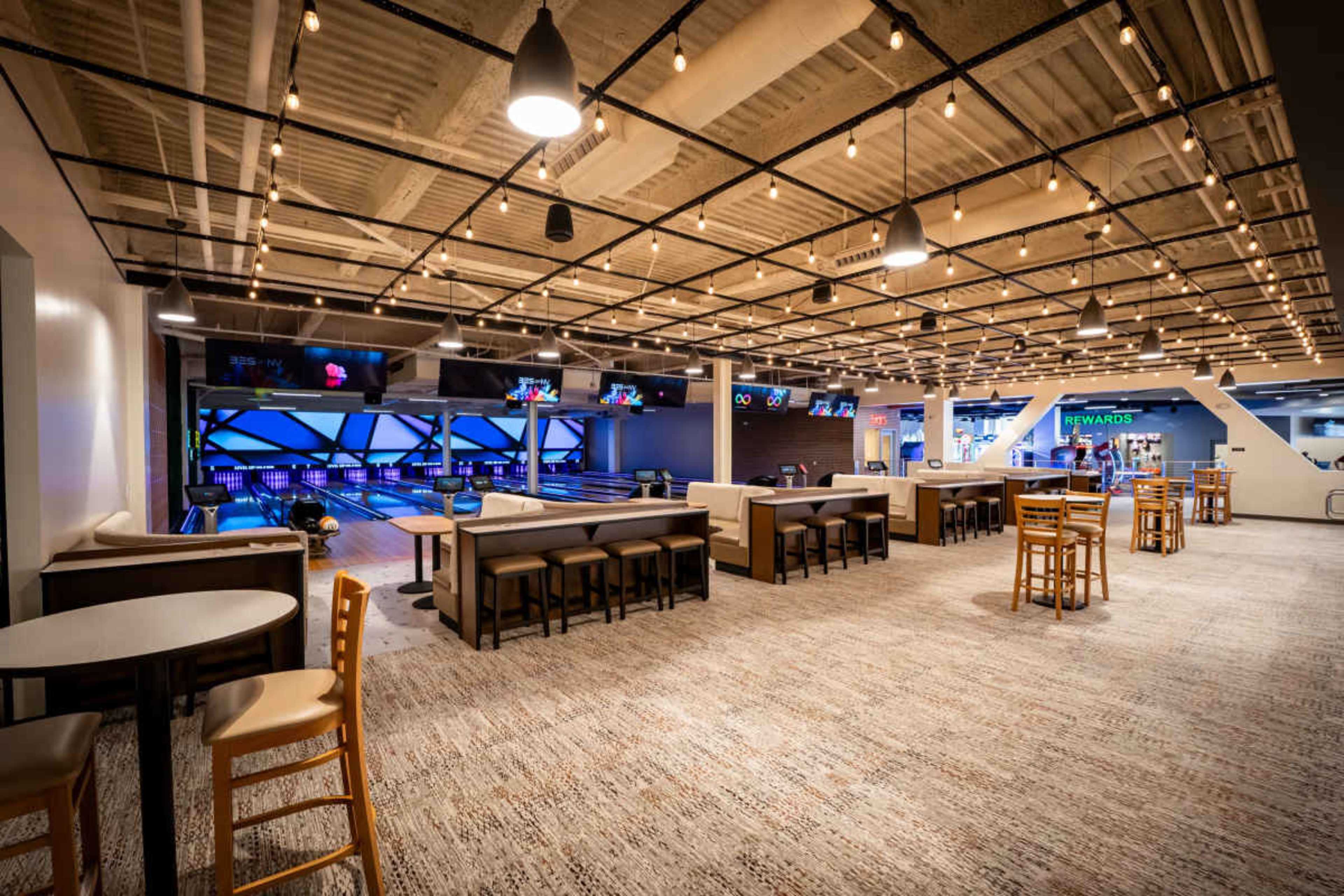 The image shows a modern bowling alley and lounge area with seating, tables, and illuminated lanes in the background.