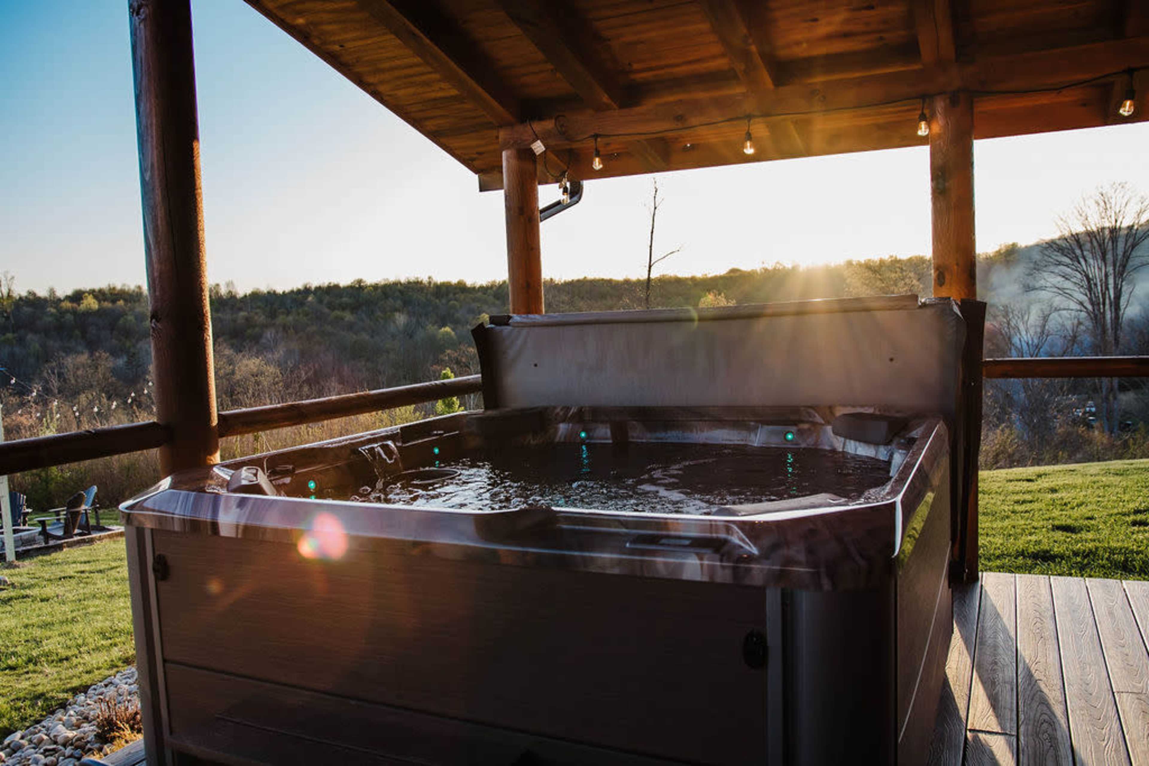 A hot tub sits on a wooden deck under a roof, with a sunset illuminating the surrounding landscape.