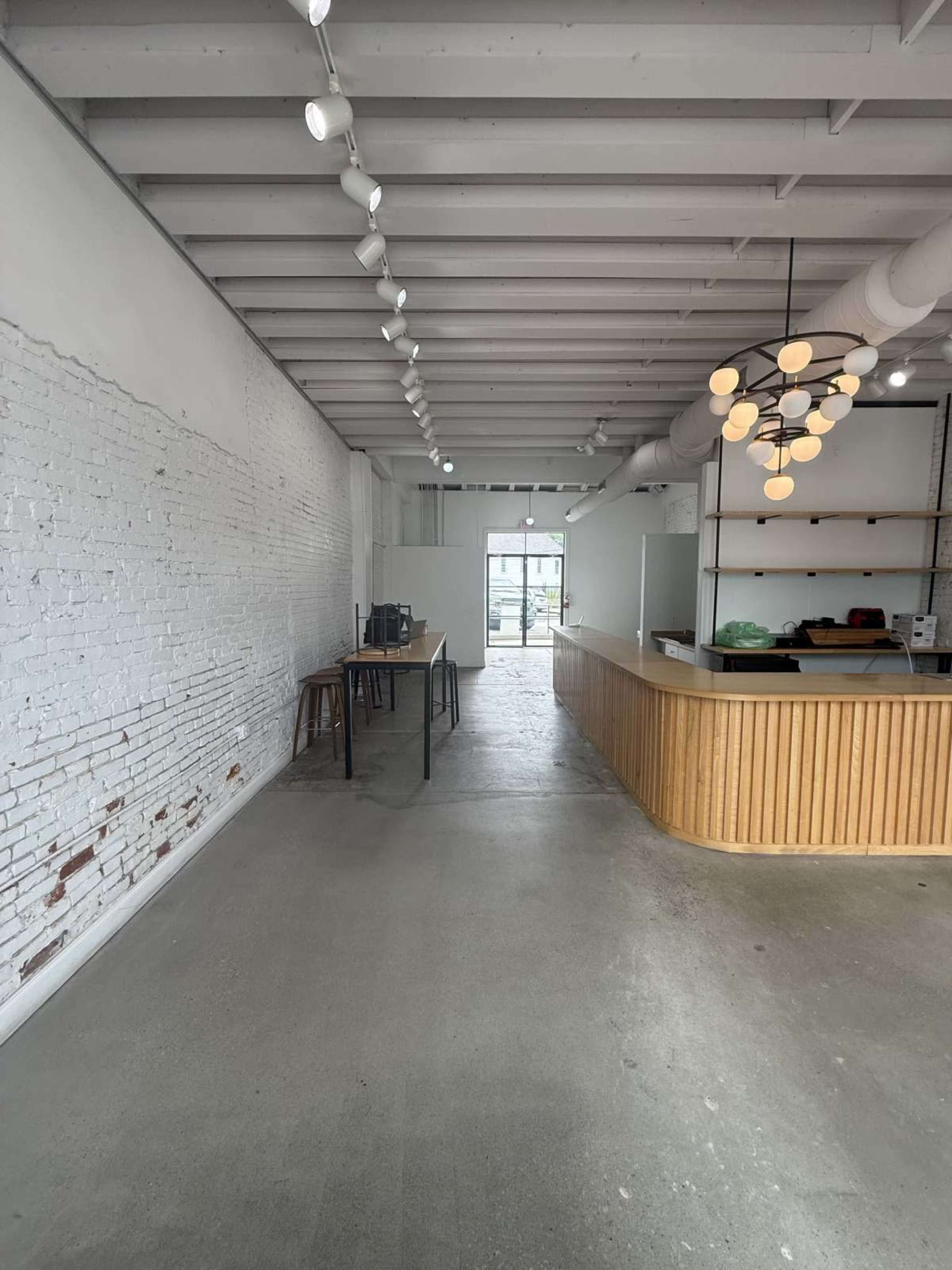 The image shows a spacious, minimally designed interior of a café with exposed brick walls, a wooden counter, and large windows at the entrance.