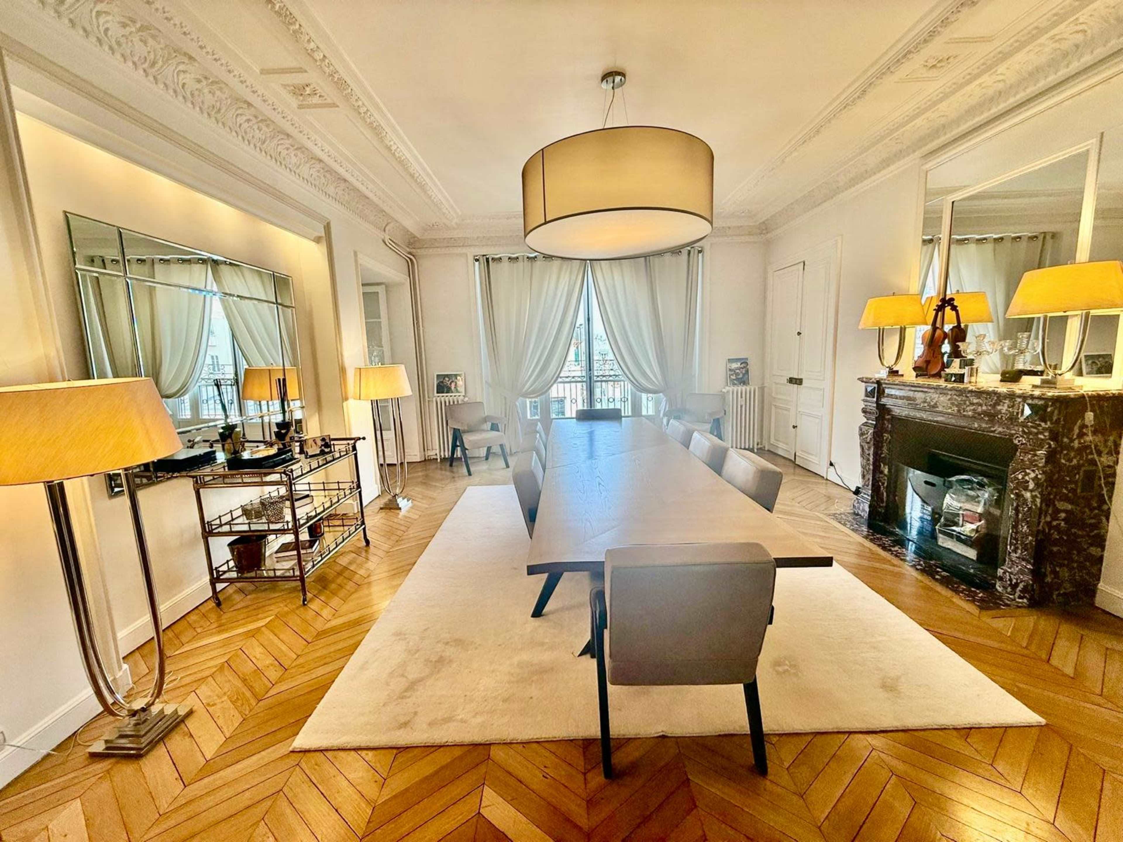 The image shows a spacious dining room with a large wooden table, modern chairs, and elegant decor, including a fireplace and two lamps.
