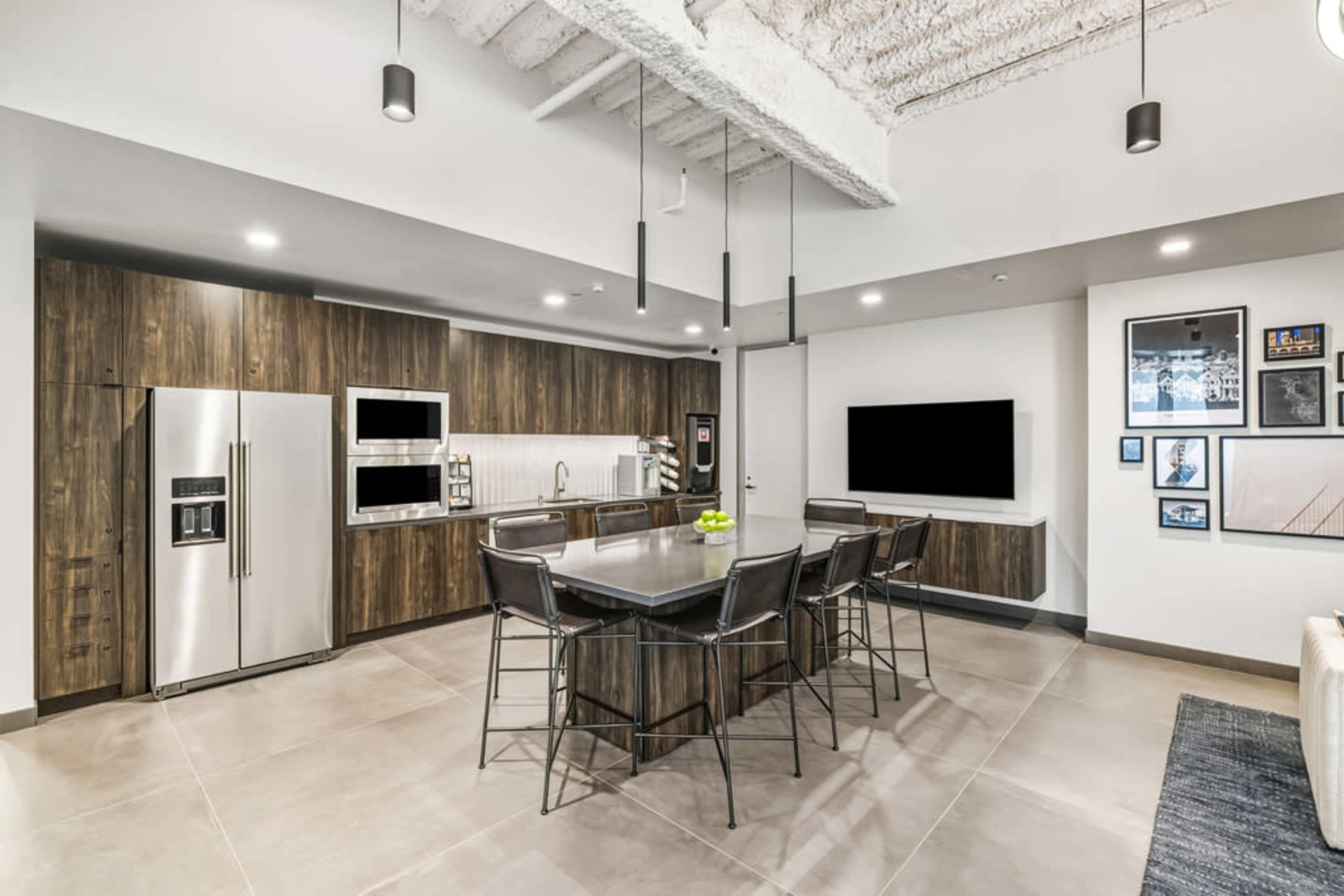 The image shows a modern kitchen with a central dining table, stainless steel appliances, and a sleek design featuring wooden cabinetry and a large flat-screen TV on the wall.