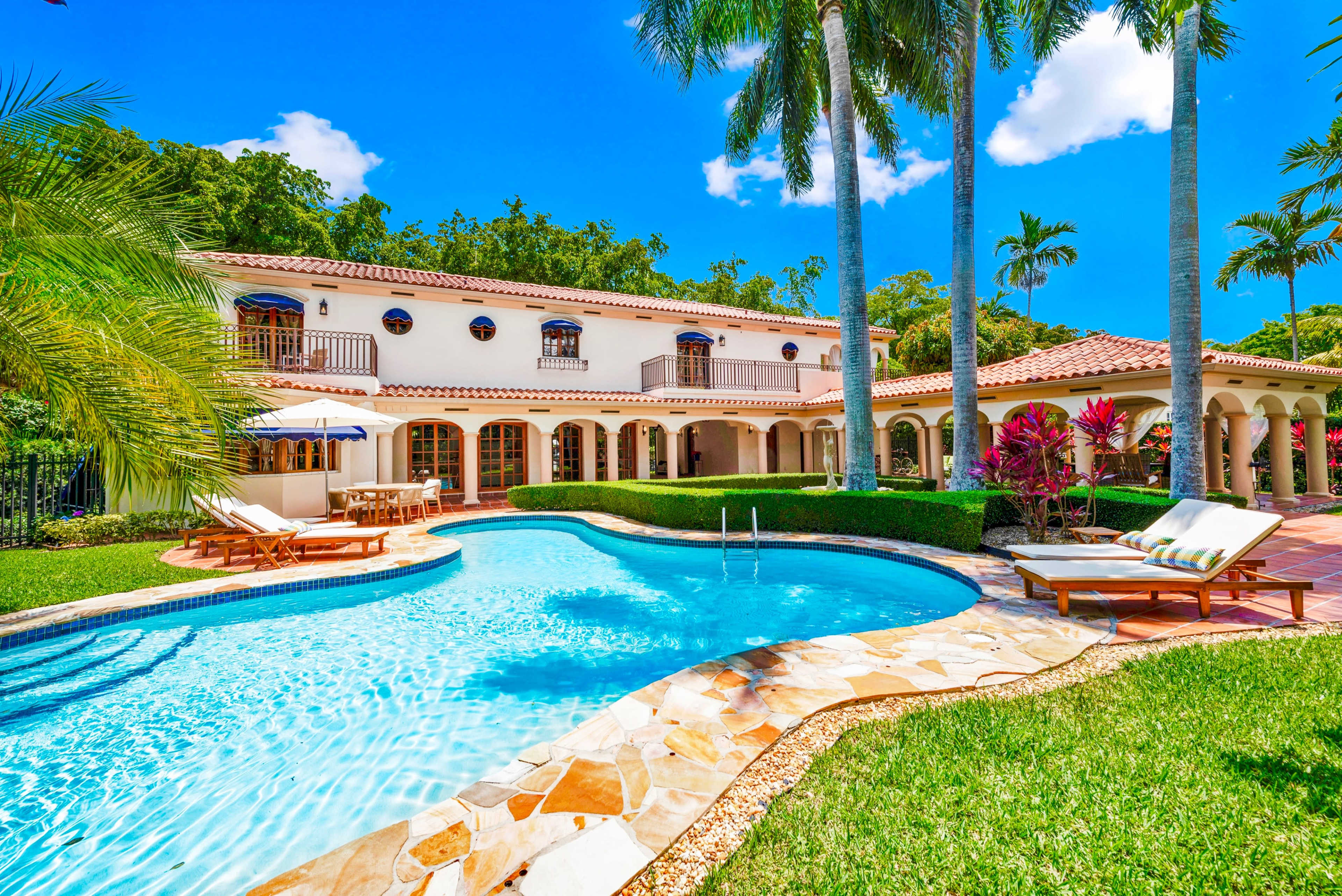 A sunny backyard featuring a swimming pool, lounge chairs, and a two-story house surrounded by palm trees.