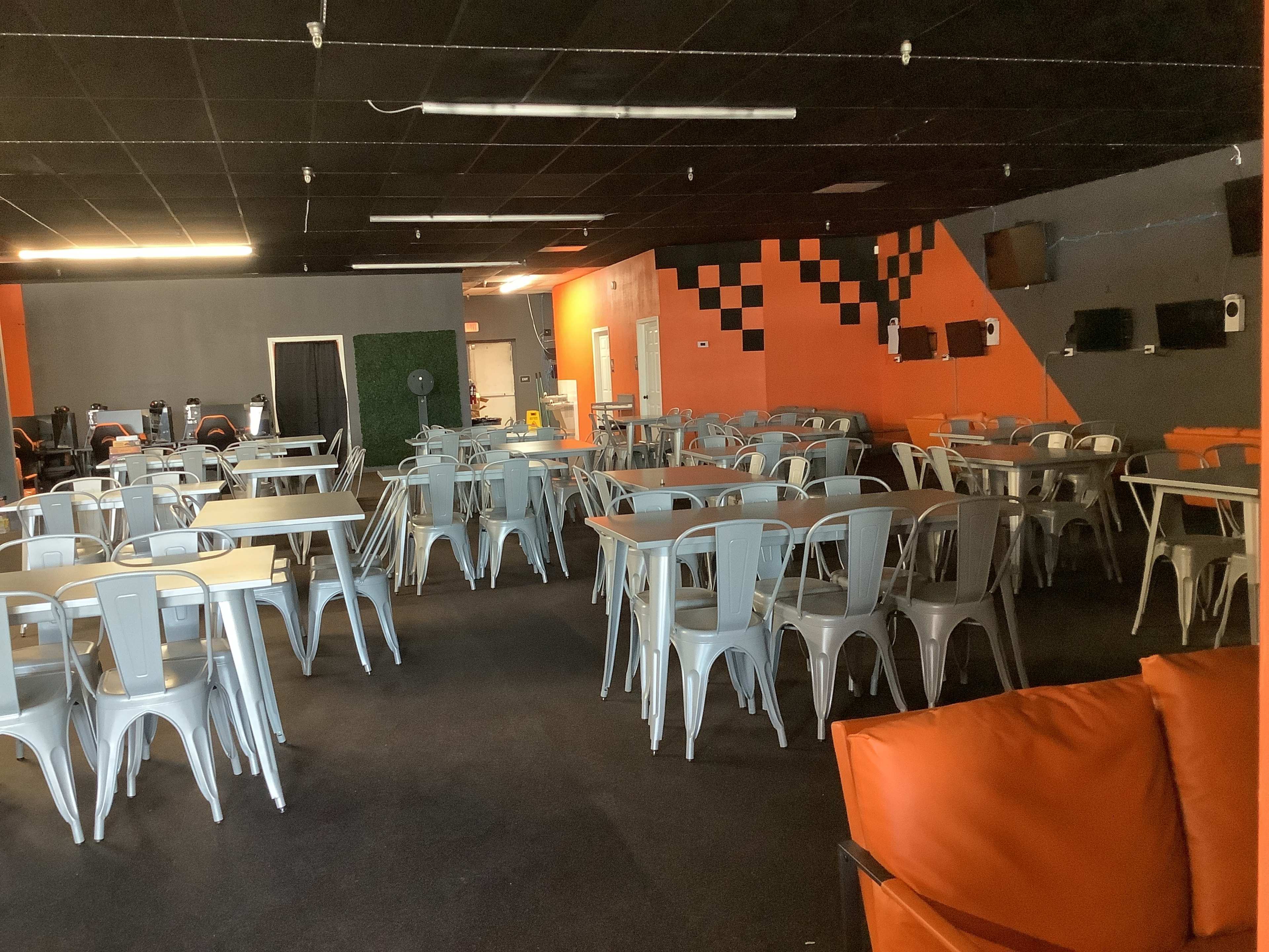 An empty dining area features numerous metal tables and chairs, with orange and gray walls and minimal decor.