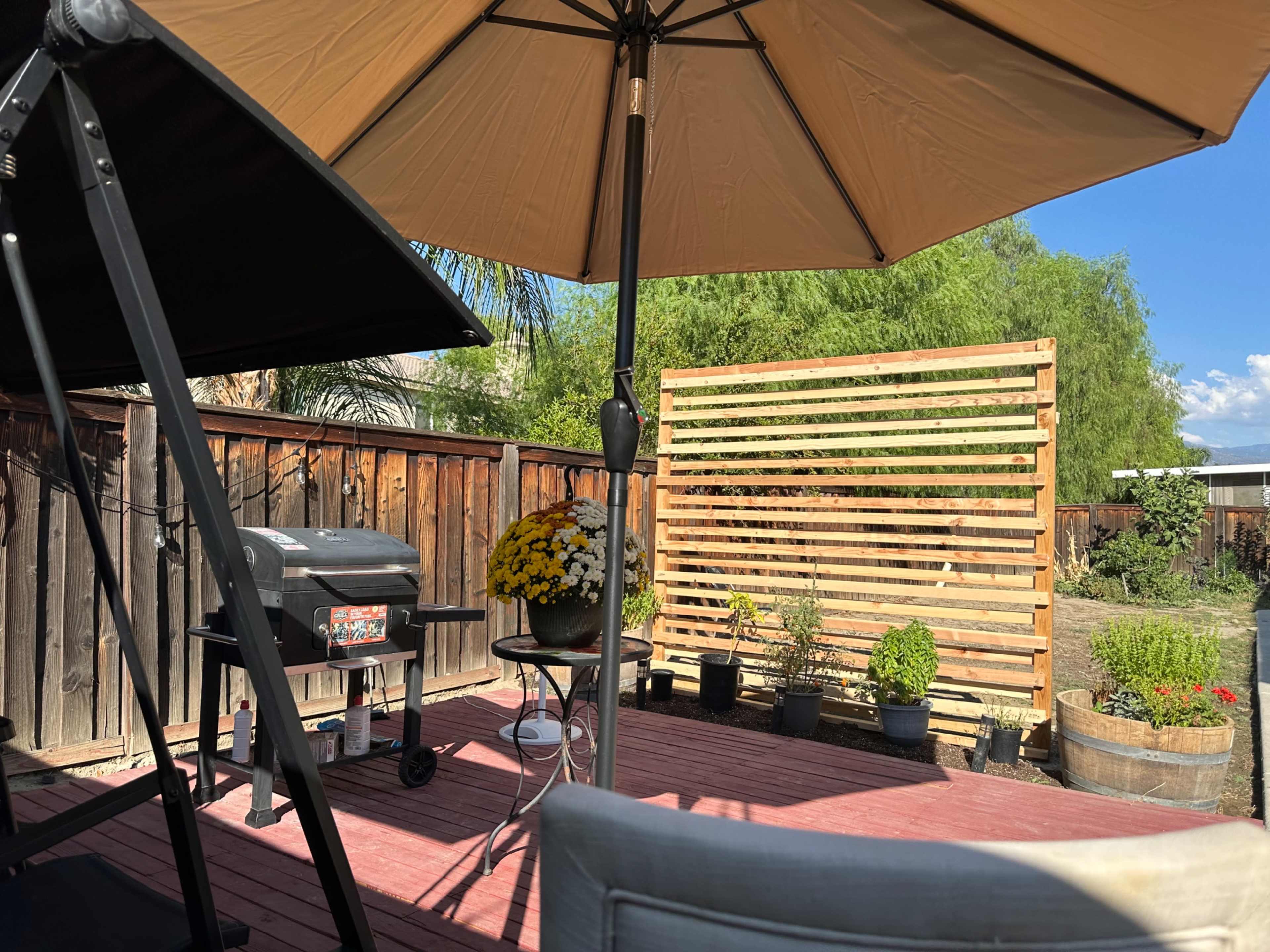 The image shows a backyard patio area with a grill, table, and potted plants under an umbrella.
