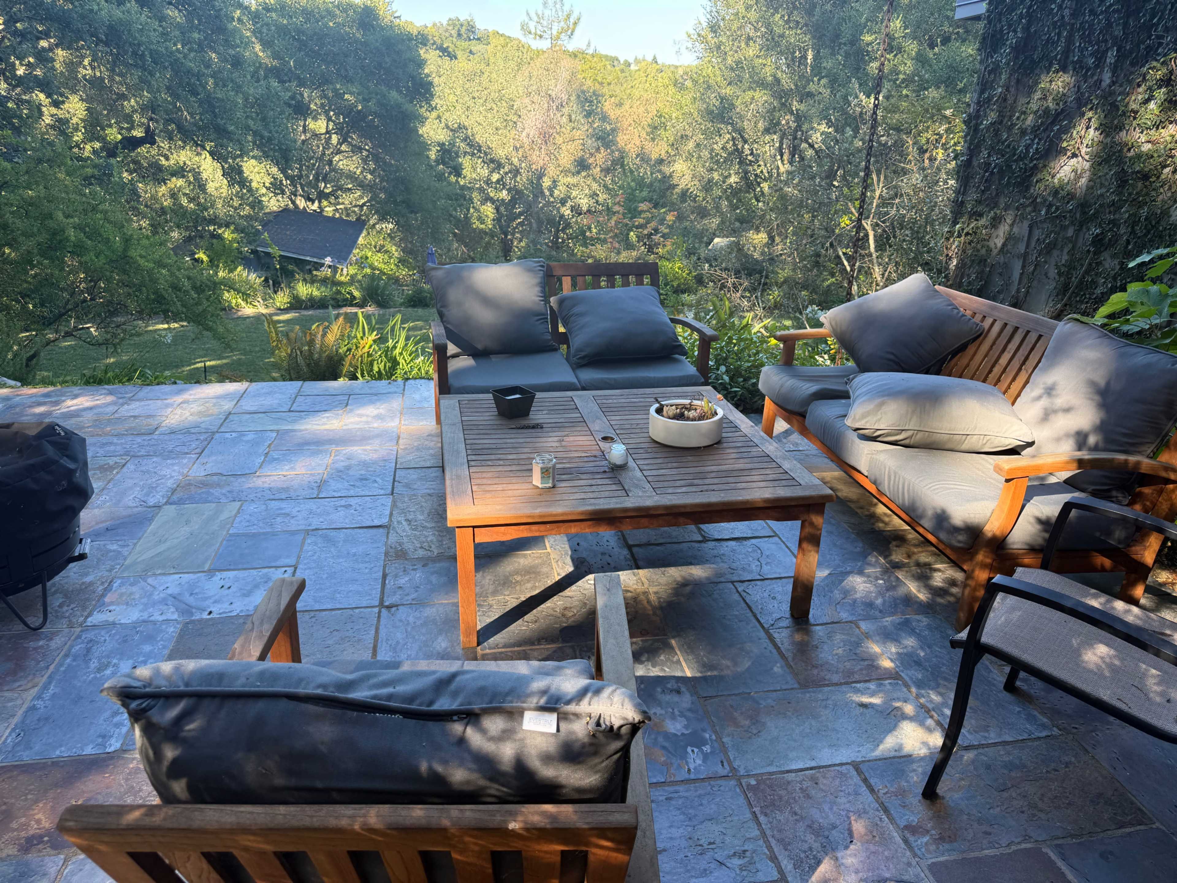 The image shows a patio area with wooden furniture, including a table and chairs, surrounded by trees and greenery.