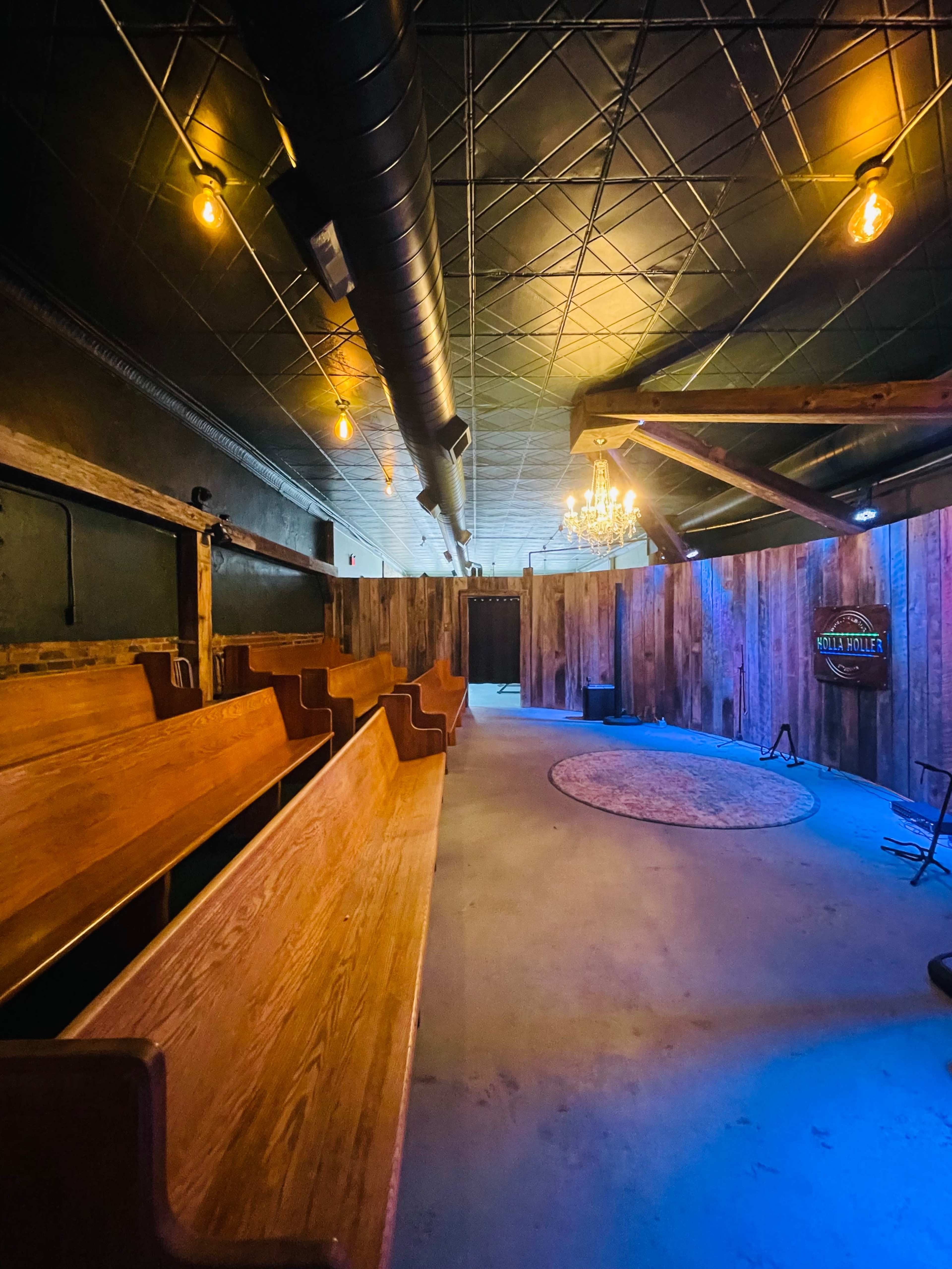 The image shows a dimly lit interior space featuring wooden pews arranged in a line, with a circular area in the center surrounded by rustic wooden walls and a chandelier.