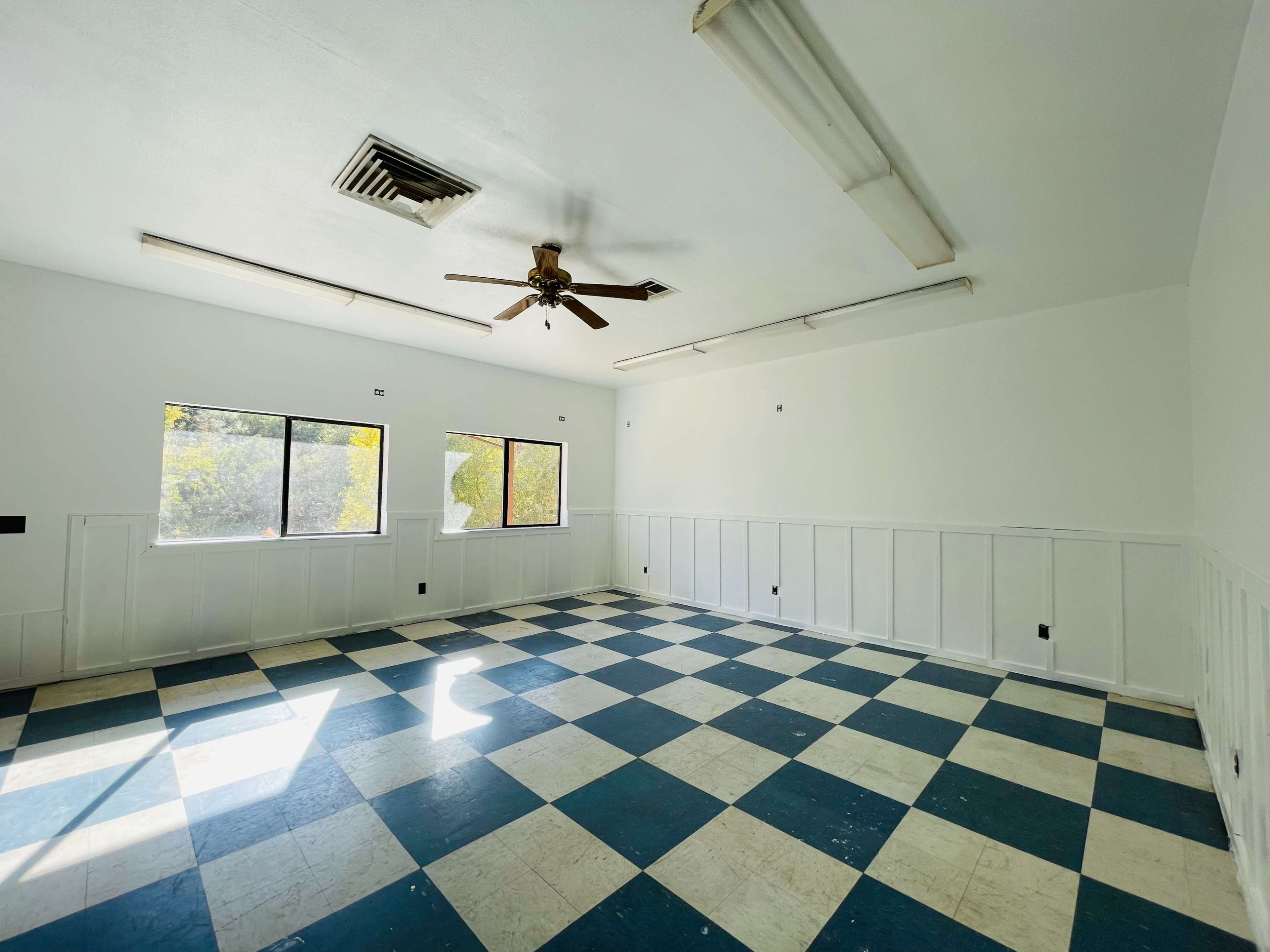 An empty room with a checkered blue and white floor, two windows, and a ceiling fan.