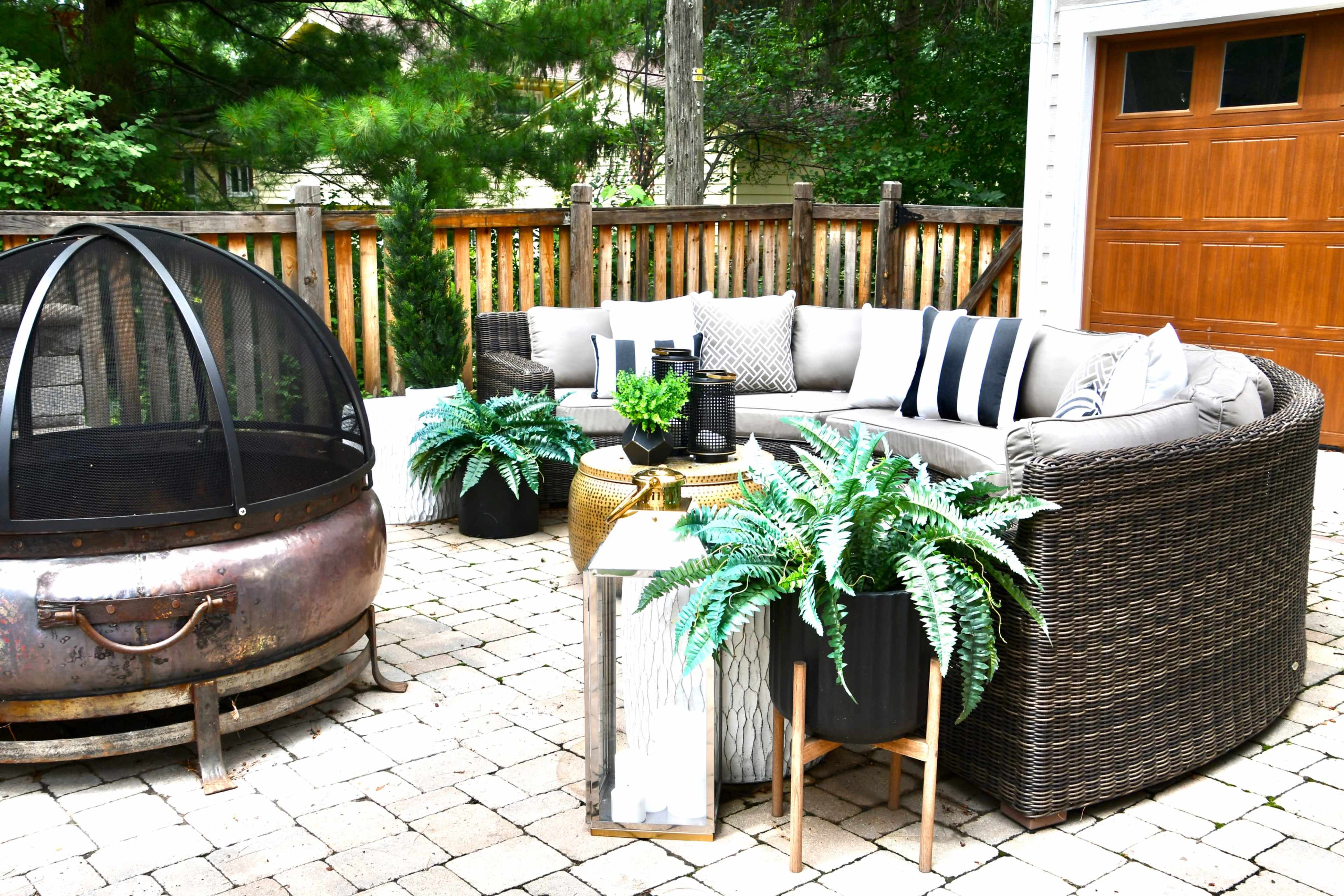 The image shows a patio area featuring a circular wicker seating arrangement, a fire pit, and potted ferns, all surrounded by a wooden fence and greenery.