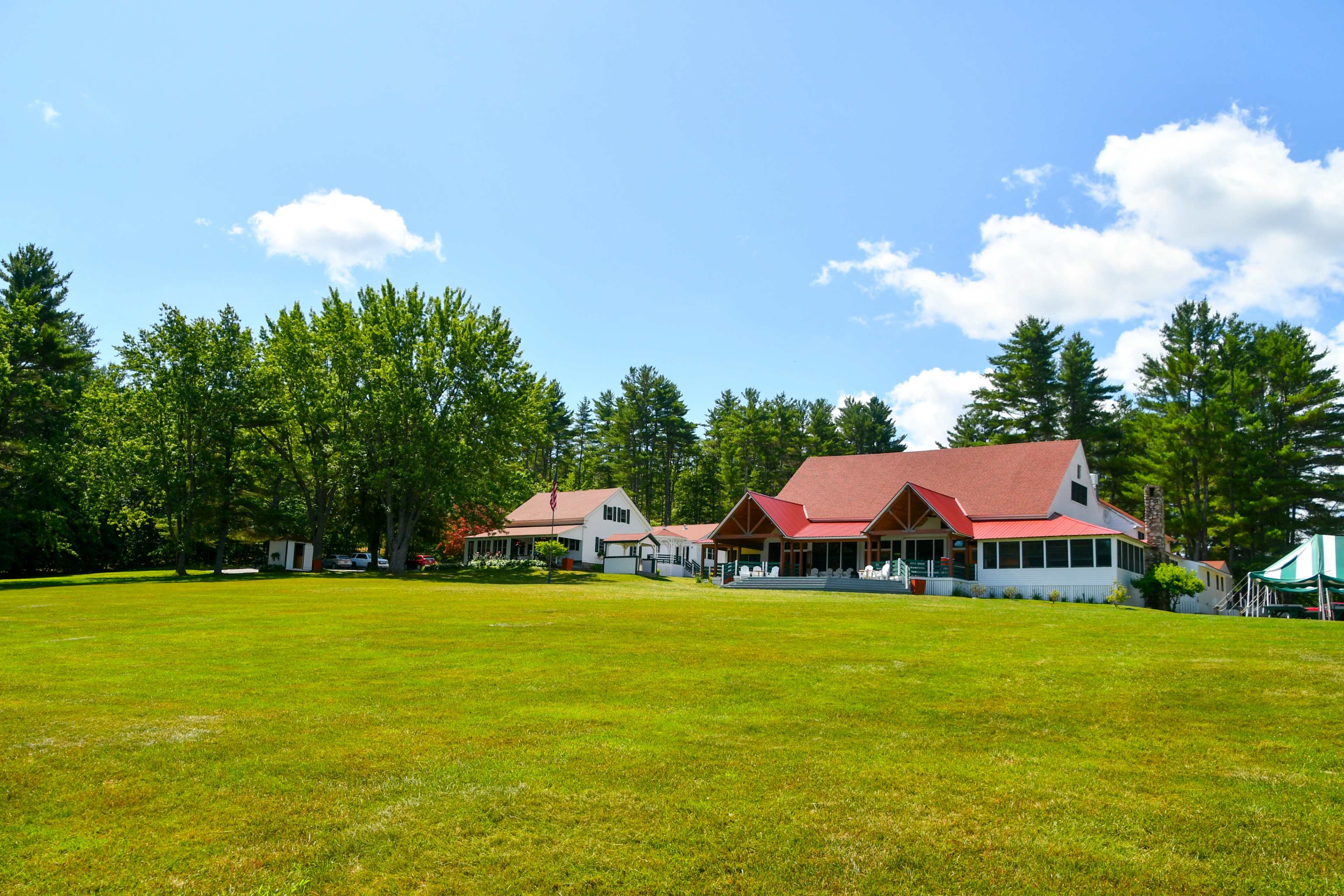 A large, open grassy area features several buildings with red roofs surrounded by tall trees under a clear blue sky.