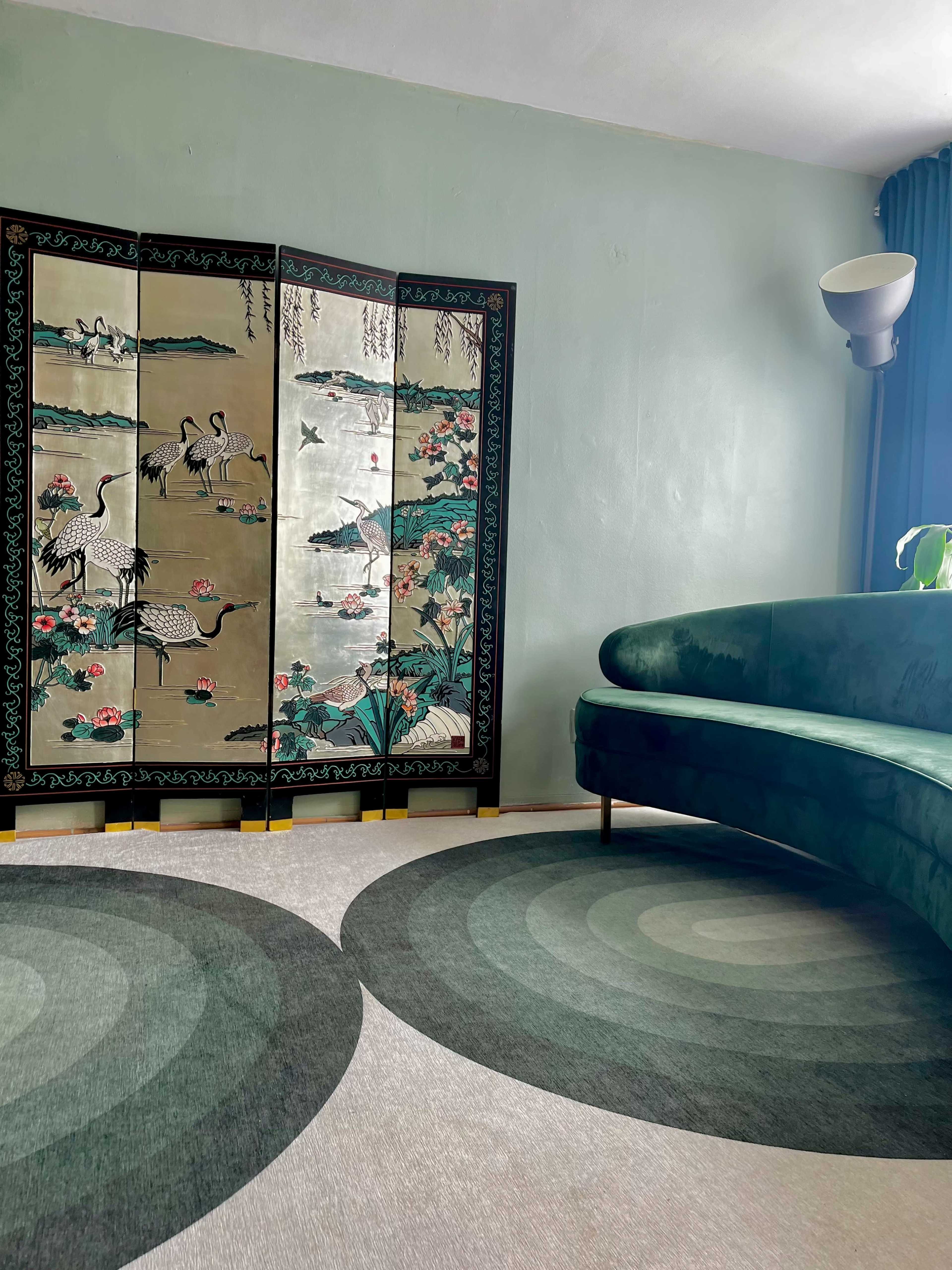 A living room corner featuring a green curved sofa, a decorative folding screen with crane motifs, and two circular rugs on a light-colored floor.