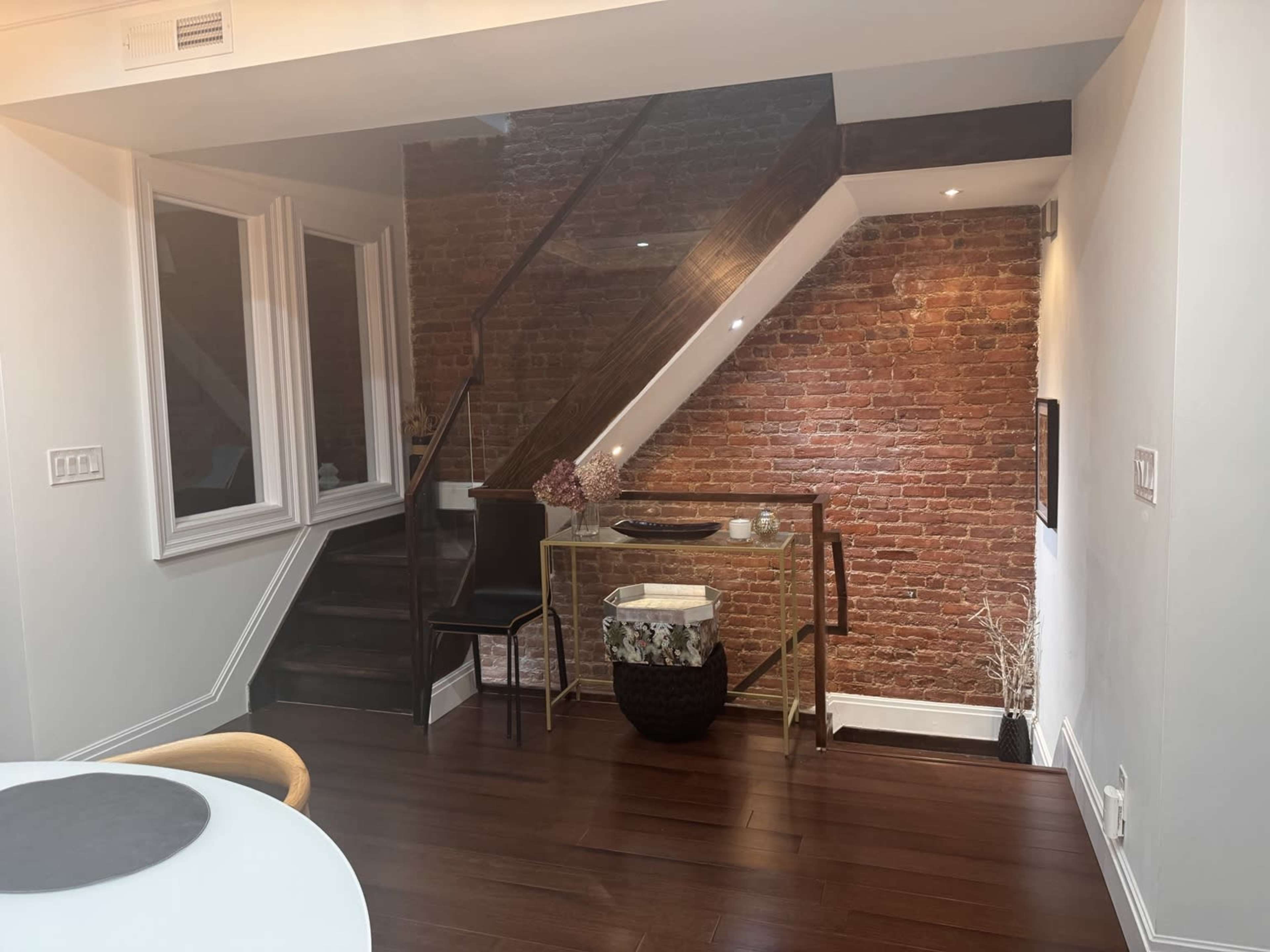 The image shows a modern interior with a wooden staircase, exposed brick wall, and a glass console table beside it.