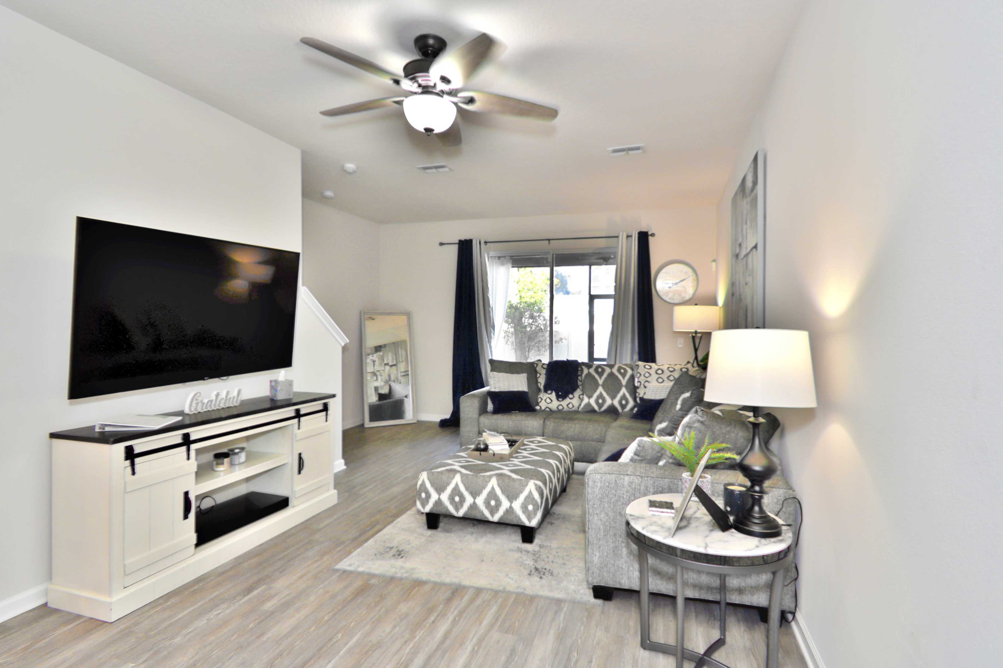 The living room features a sectional couch, a large flat-screen TV mounted on the wall, and a lamp beside a round table, creating a cozy and modern space.