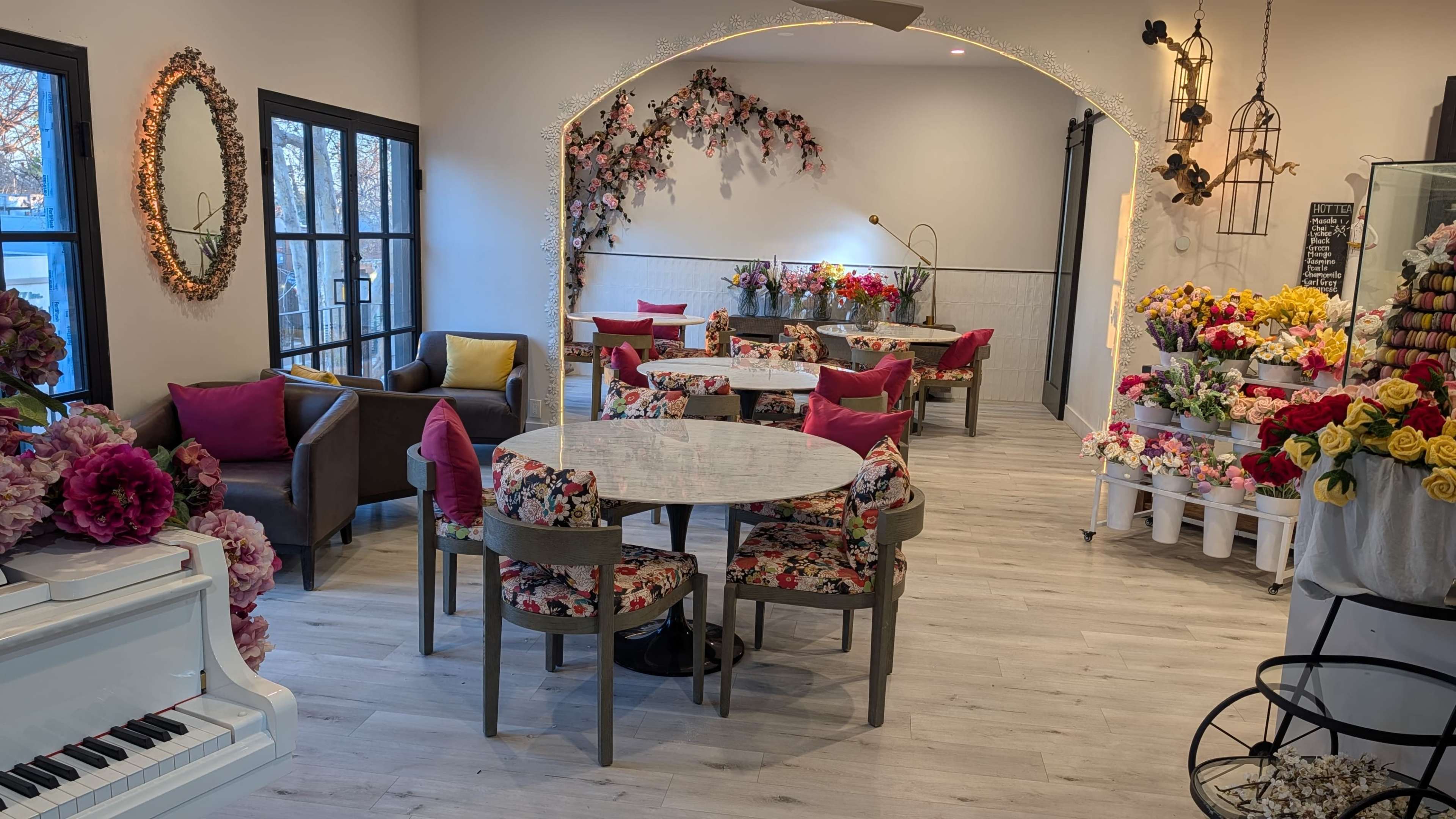 The image shows a brightly lit café featuring floral-themed furniture, a white piano, and a display of colorful flowers.