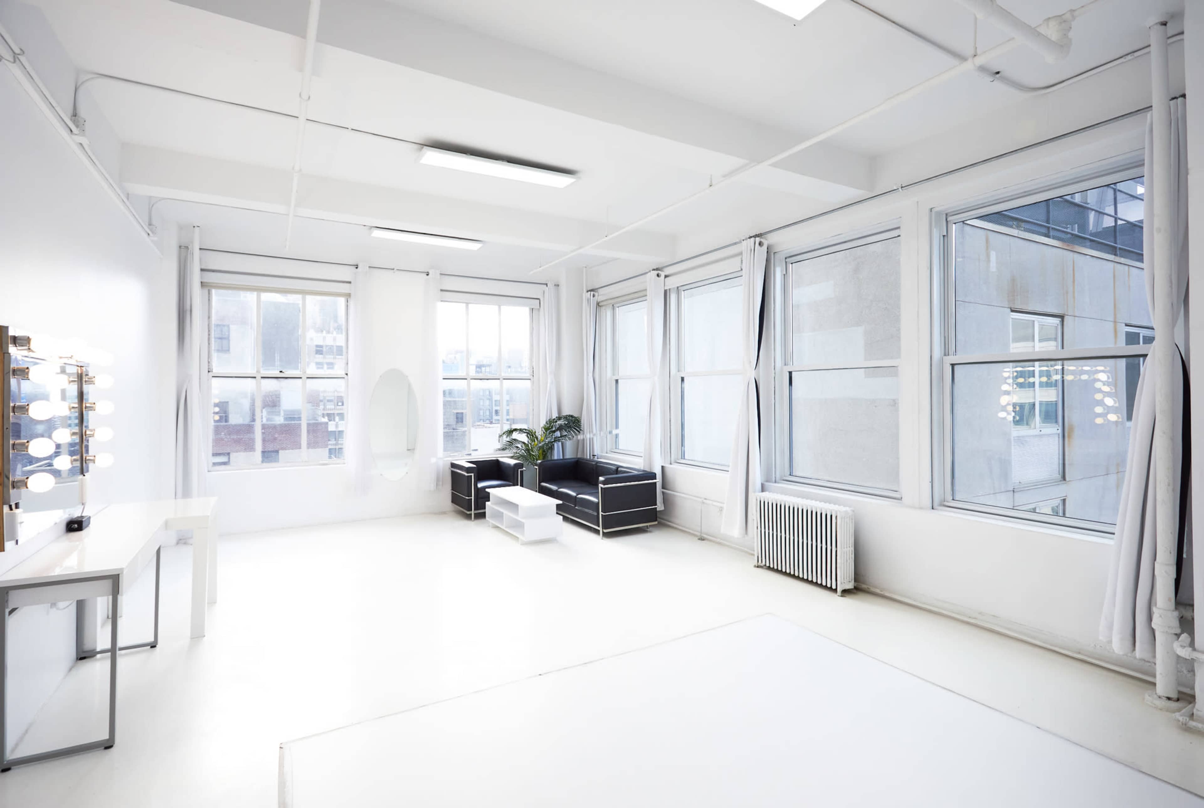The image shows a bright, spacious room with large windows, light-colored walls, and minimal furniture including a vanity mirror and a small seating area.